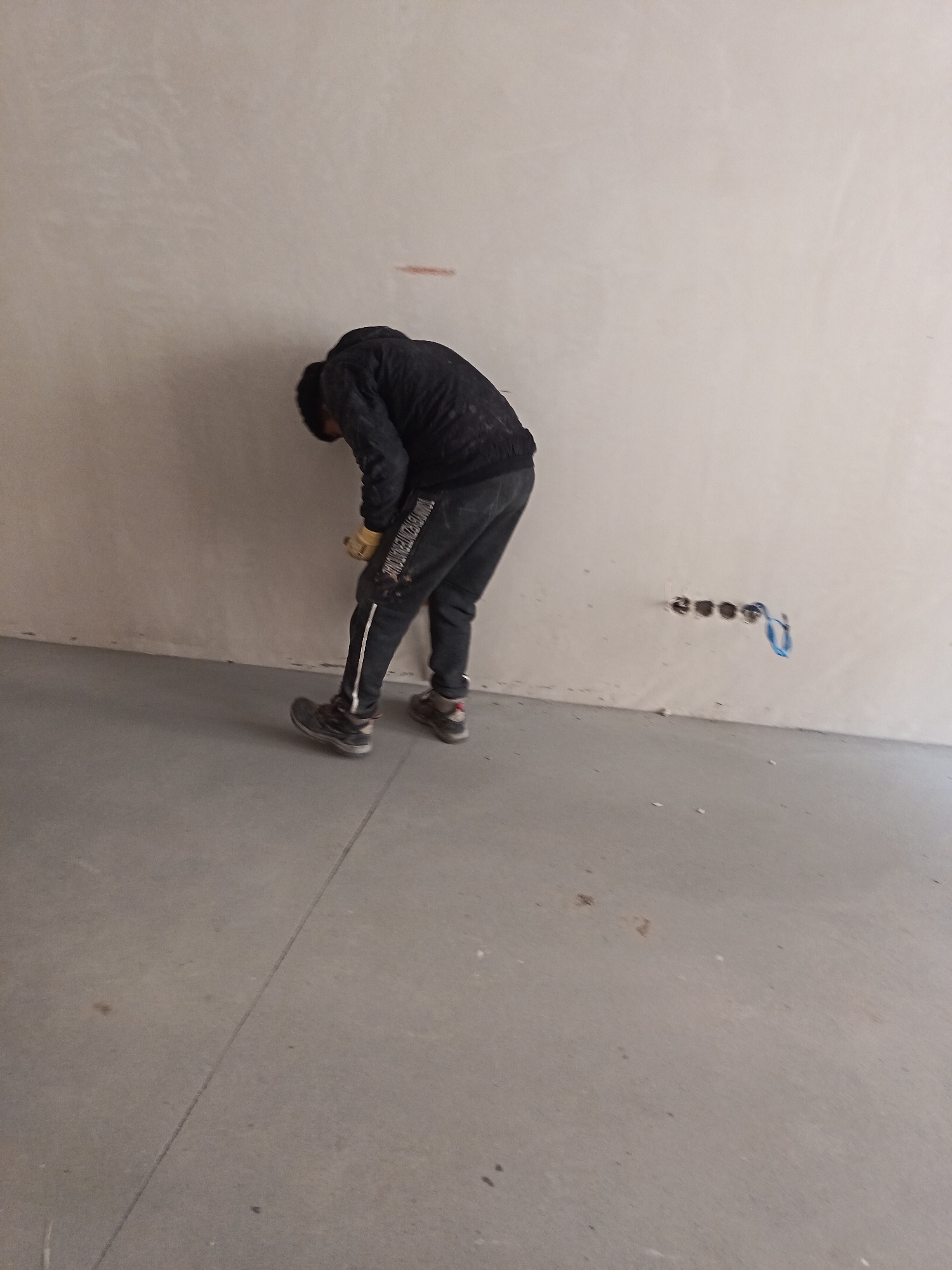 An interior scene in Sofia, Bulgaria, depicts a single individual engaged in work within an unfinished or renovated room. The person, positioned in the center-left, is bent forward at the waist, facing a light-colored wall. They wear a dark long-sleeved top, dark athletic trousers with white vertical stripes and partially legible text "TOMMY HILFIGER INTERNATIONAL" on the left leg, light-colored work gloves, and dark athletic shoes. Their gloved hands are near the lower portion of the wall. The wall itself is light-colored with minor surface variations; a faint, horizontal reddish mark is visible on the upper wall section. To the right, four circular openings, presumably for electrical outlets or conduits, are horizontally aligned in the wall, with a blue electrical wire extending from the rightmost opening. The floor is a light grey concrete or screed surface, featuring a visible straight seam running across it in the mid-ground, and scattered minor debris.