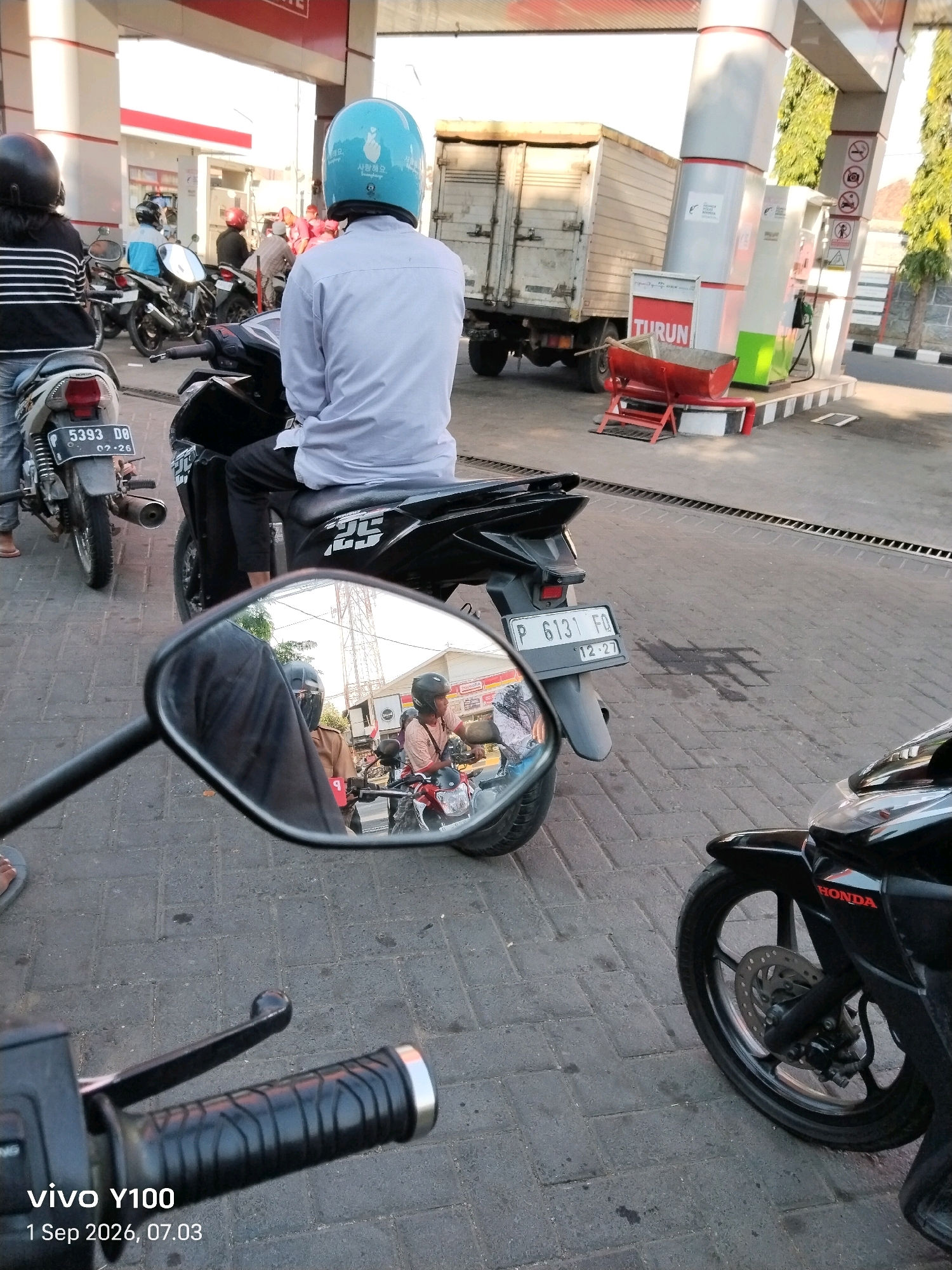 The image depicts a scene at a gas station in Situbondo, Indonesia, on September 1, 2026, at 07:03 AM. The foreground shows the handlebars of a black motorcycle, with a timestamp overlay reading "vivo Y100 1 Sep 2026, 07.03". In the mid-ground, a person wearing a blue helmet is seated on a black motorcycle. The helmet has Korean writing and a hand gesture symbol on it. This person is wearing a light blue long-sleeved shirt and dark pants. Behind them, another motorcycle with a license plate reading "P 6131 FQ 12-27" is visible. A side-view mirror attached to a motorcycle in the foreground reflects another motorcycle with a rider wearing a brown uniform and helmet. In the background, a large white truck is parked near gas pumps. Several other motorcycles and people are present at the gas station. The ground is paved with interlocking bricks. The environment appears to be an outdoor setting, with trees and the roof of the gas station structure visible.