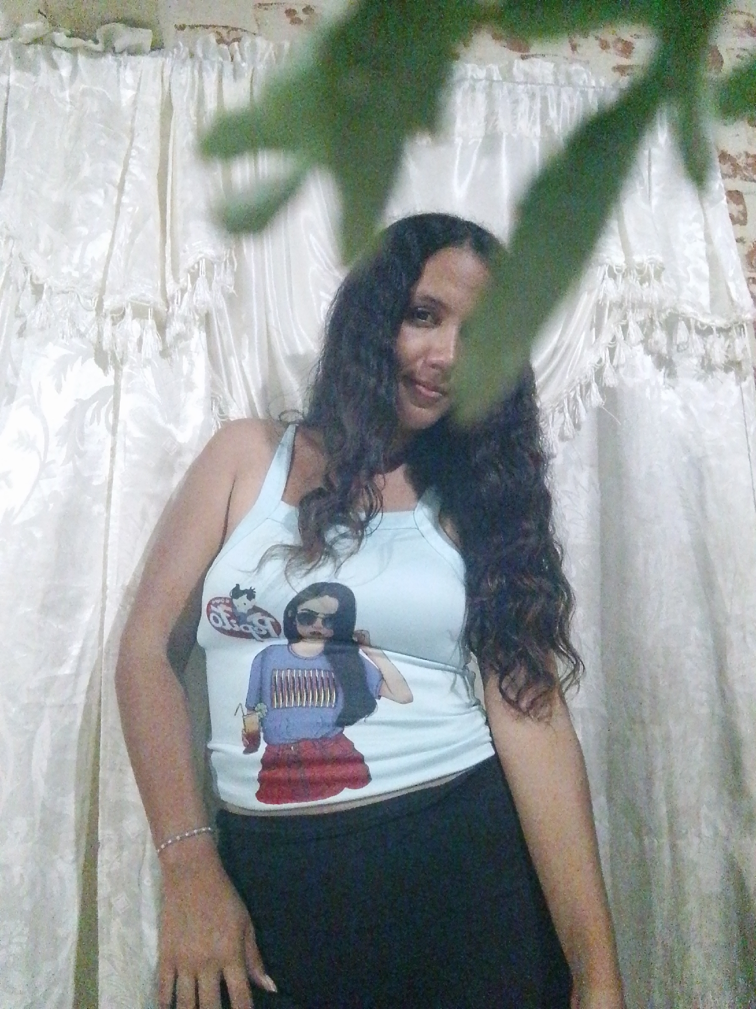 A woman stands in front of a white curtain with a patterned valance, partially obscured by out-of-focus green foliage at the top of the frame. The woman has long, dark, wavy hair and is wearing a light blue tank top with a graphic design featuring a female figure and a cartoon character. She is also wearing dark pants. A delicate bracelet is visible on her left wrist. The background shows a wall with a slightly textured surface, possibly brick or plaster. The location is Juan Griego, Venezuela.