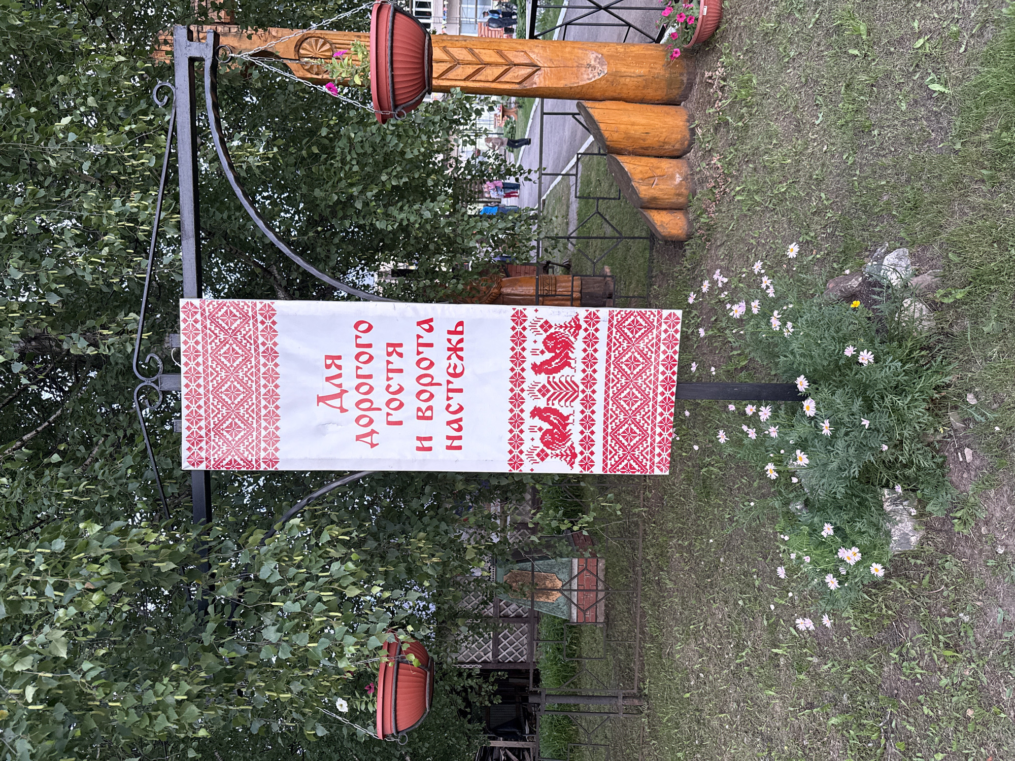In the city of Kukui, Russia, a decorative banner hangs from a black metal archway, flanked by birch trees. The banner, primarily white with red embroidered details, features Cyrillic text that reads "Для дорогого гостя и ворота настежь" (For a dear guest, the gates are wide open). Below the text, two red roosters are embroidered, followed by intricate geometric patterns. Two red hanging planters, one with a white flower and another with pink flowers, are suspended from the archway. A wooden post with carved details stands to the right of the banner. The foreground is a grassy area with scattered daisies and some rocks. In the background, a fence encloses a yard with a checkered fence and a small monument. A pathway is visible, with several people walking in the distance.