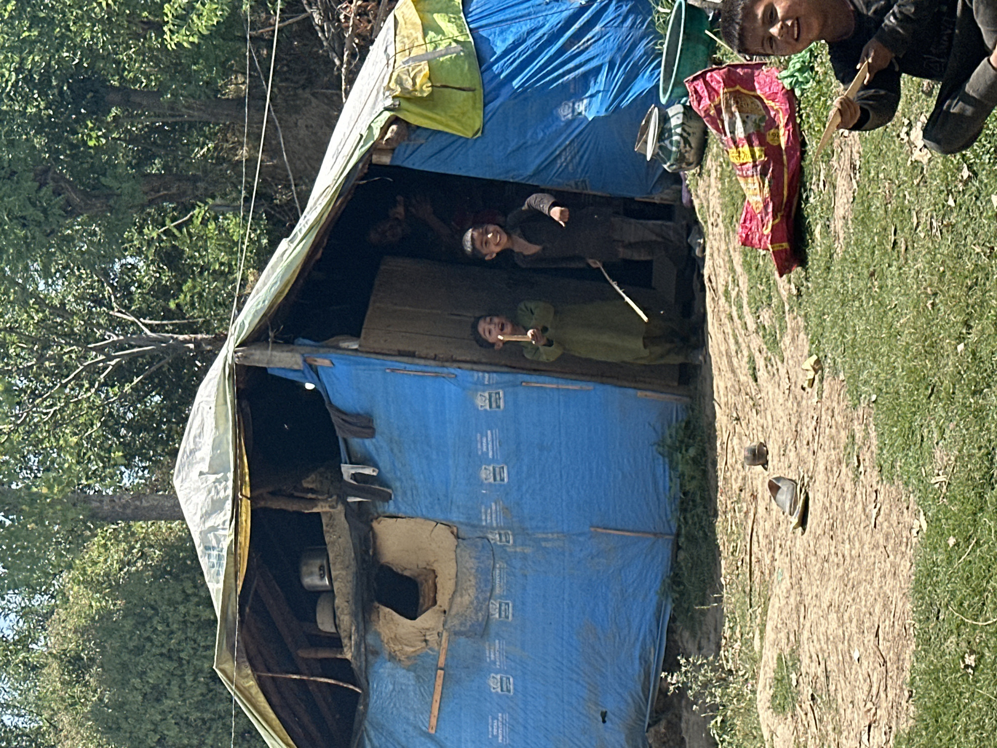 A group of children are playing in front of a makeshift shelter made of blue tarpaulin and a patched roof. The shelter appears to be in a rural or impoverished area, with dirt ground and overgrown grass. Inside the shelter, cooking pots are visible. One child in the foreground, looking directly at the camera, is holding a stick. Two other children are standing in the entrance of the shelter, one holding a stick as well. A bright red and yellow object, possibly a blanket or mat, lies on the ground near the shelter. The background consists of trees and foliage, suggesting an outdoor setting.