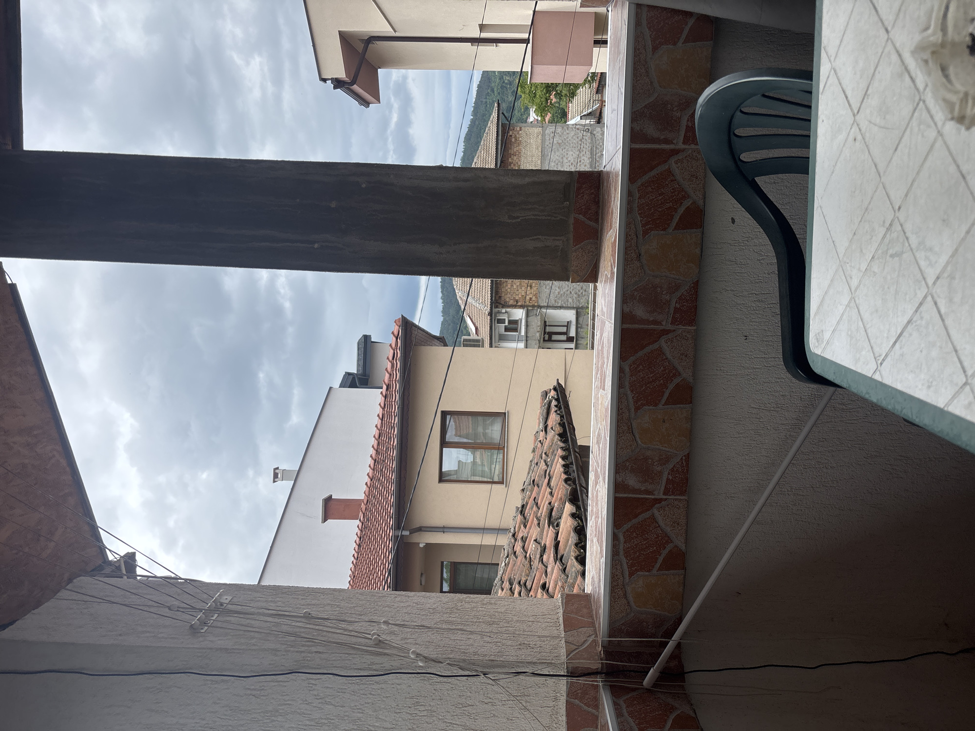 The image is taken from a balcony or porch in Asenovgrad, Bulgaria, looking out between buildings. The sky is overcast. A concrete pillar and the edge of a building frame the view. Buildings of varying heights and architectural styles are visible, with red tiled roofs and beige or white walls. In the distance, tree-covered hills are visible. Part of a plastic chair and a tiled table are in the foreground on the right side of the image. Power lines are visible stretching across the lower portion of the frame. The overall impression is of a residential area in a small city.