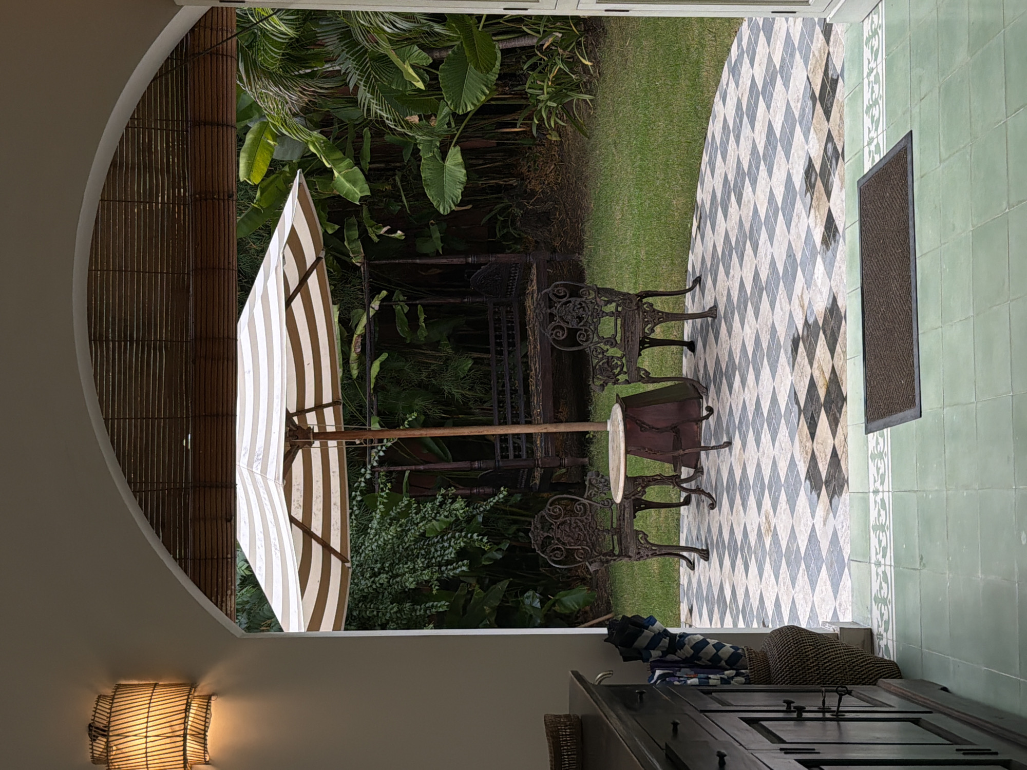 The image shows an open doorway looking out onto a lush green garden area. In the foreground, there is a dark wooden cabinet with several drawers and doors, and to its left, a woven basket and an umbrella stand with blue and white striped umbrellas. The floor is tiled with a pattern of green tiles leading to a checkered tiled patio. On the patio, there is a round table with two ornate cast-iron chairs. Behind the patio, a large striped umbrella shades a seating area. Further back in the garden, a dark wooden swing set or daybed is visible amongst dense tropical foliage. The scene appears to be a tranquil outdoor living space, possibly a resort or private residence in North Kuta, Indonesia, given the tropical vegetation. There are no people present in the image.
