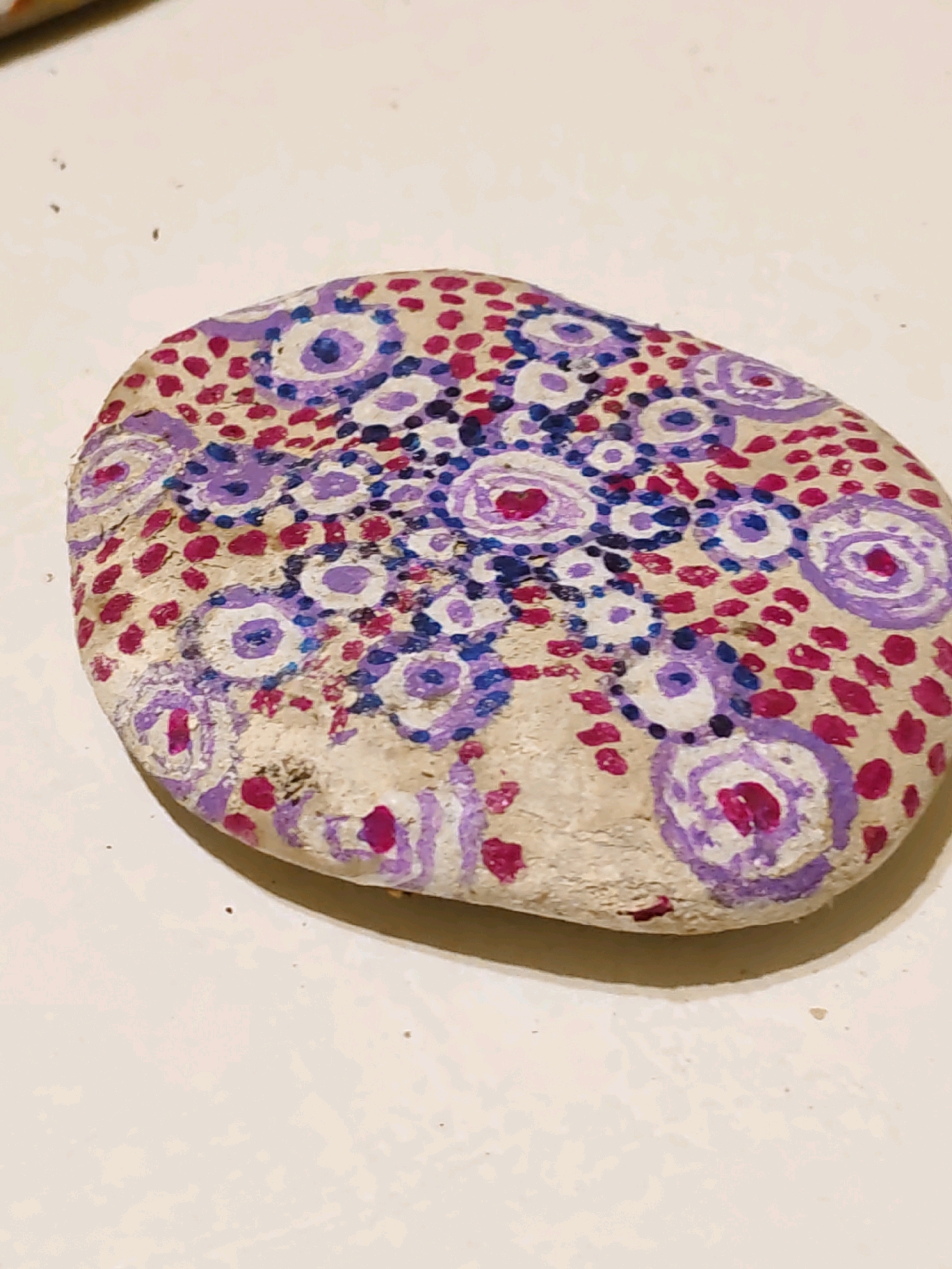 The image shows a close-up, overhead view of a painted, oval-shaped stone resting on a light-colored, textured surface. The stone is decorated with a pattern of colorful dots and concentric circles. The dots are primarily magenta and dark blue, scattered across the surface. The concentric circles are painted in purple, white, and magenta, with some having a magenta center. The stone appears to be rough and natural, with its original texture visible beneath the paint. The lighting suggests an indoor environment, possibly a table or countertop. There is no indication of Rostov-on-Don, Russia, or any specific location in the image.