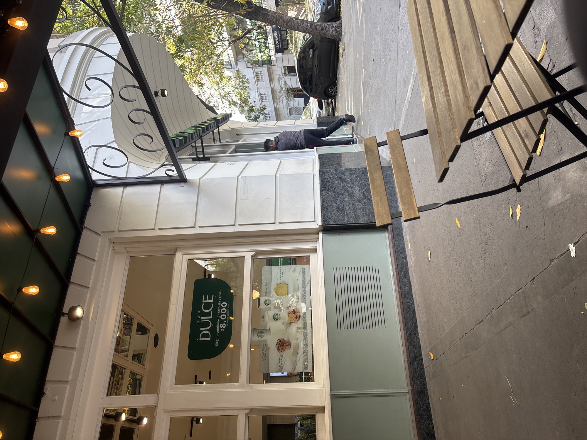 This media file captures an outdoor urban scene featuring a building facade, a person, and street furniture under natural daylight. The image is taken from a slightly upward angle, showing a storefront with an ornate awning and outdoor seating.
The main subject is a male figure, seen from the back, leaning against the white facade of the building. He is wearing dark trousers and a patterned grey long-sleeved top. To the right, a wooden slatted table and two matching chairs are set up on the grey pavement, indicative of a sidewalk cafe or restaurant. The ground is a paved sidewalk with scattered yellow leaves.
The building itself has a white, paneled facade with large windows. A dark green awning with decorative black wrought-iron scrollwork extends over the entrance. String lights with warm-toned bulbs are visible beneath this awning. Through the large front window, a promotional poster is displayed, advertising "DUO DULCE" (Sweet Duo) with the text "Elegi tu combinación favorita por solo $8.000" (Choose your favorite combination for only $8,000). The poster also shows images of various beverages and pastries, including "Vainilla Cream Frappuccino," "Latte," "Café del Dia," "Lemon Pound," and "Muffin Choco Chip." Another partially visible text reads "OS NACIONALES SE 612."
In the background, beyond the person and the cafe, a street with a dark car parked under a leafy tree is visible. Further down the street, another building can be seen with the text "LA COVACINA" on its facade. The bright sunlight creates distinct shadows, suggesting a clear, sunny day. The currency "$8.000" (likely Argentine Pesos) along with the Spanish text confirm the location as Buenos Aires, Argentina.