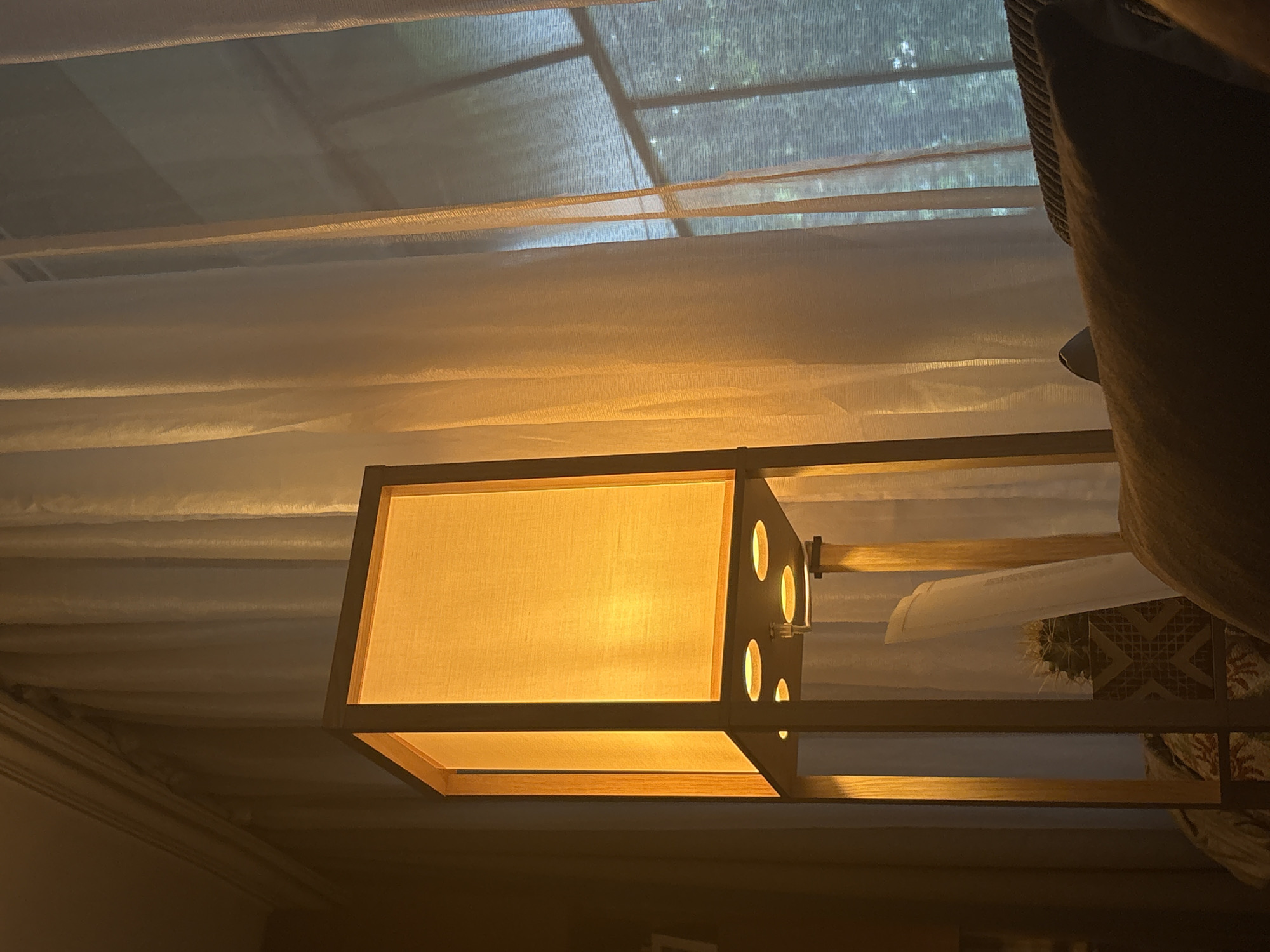 A tall, rectangular floor lamp with a wooden frame and an orange fabric shade is lit from within, casting a warm glow. The lamp stands in what appears to be a living room, with sheer white curtains covering a window behind it. Through the curtains, the faint outline of trees is visible, suggesting an outdoor setting. In the foreground, a portion of a dark-colored sofa is visible, with a patterned throw blanket draped over it. A potted cactus plant sits on a lower shelf of the lamp. The image is taken from a low angle, looking up towards the lamp and window. The overall atmosphere is cozy and dimly lit, with the lamp providing the primary source of illumination. The location is not discernible from the image and therefore cannot be identified as Sofia, Bulgaria.