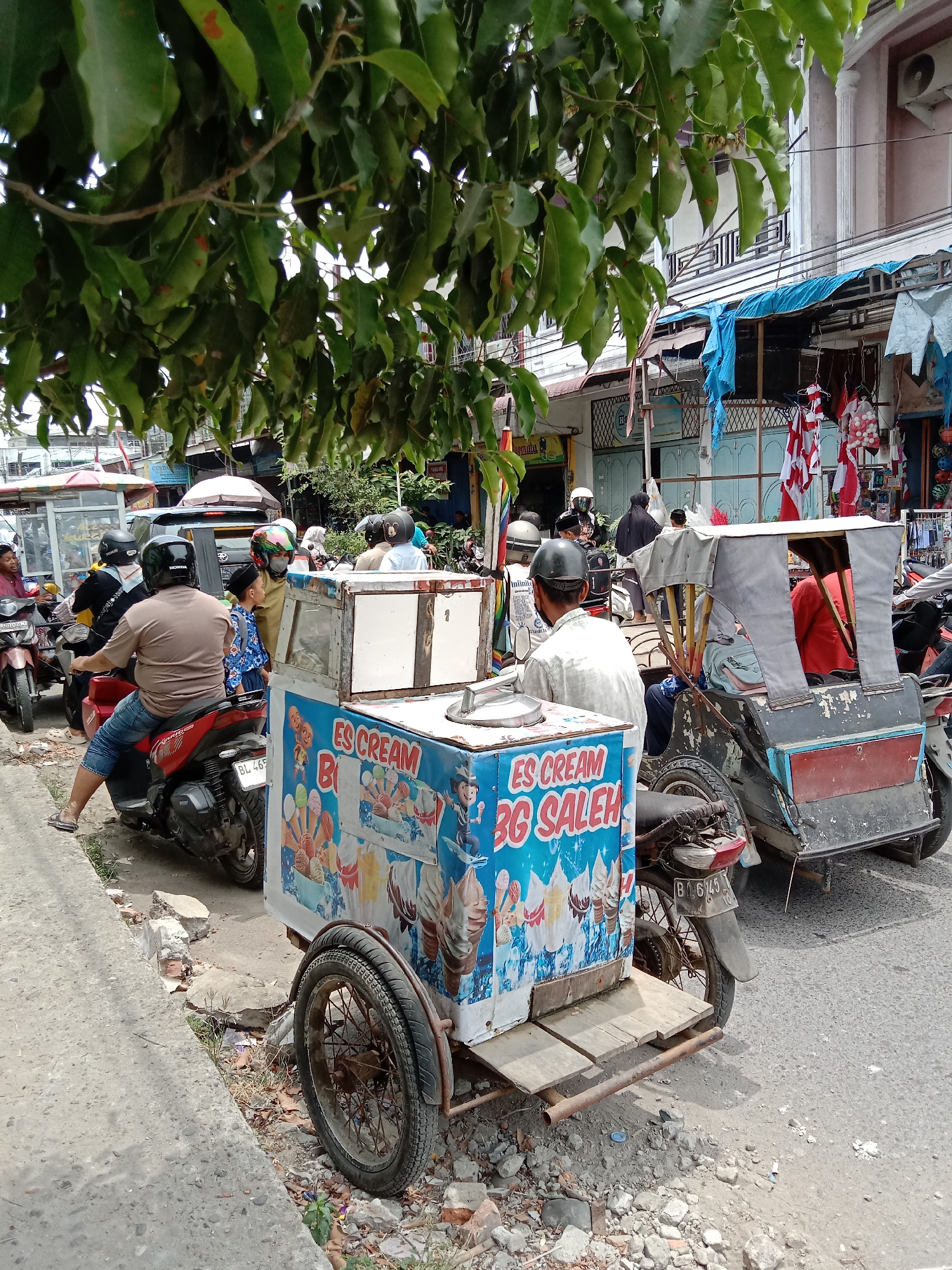 A street scene in Lhokseumawe, Indonesia, features an ice cream cart on the side of the road, partially obscuring a rickshaw. The ice cream cart is decorated with images of ice cream cones and has "ES CREAM BG SALEH" written on its side. The rickshaw, a three-wheeled vehicle, is weathered and has a grey fabric cover. Several people are visible on motorcycles, suggesting traffic or a busy marketplace. One person is seated on a red motorcycle, facing away from the camera. Another person, wearing a helmet, is standing near the ice cream cart. The background shows buildings with shops and awnings, typical of a commercial street. Trees with green leaves overhang the scene from the top, casting shadows. The road surface is asphalt, with debris and some vegetation along the roadside. The overall impression is of a bustling urban environment in Indonesia.