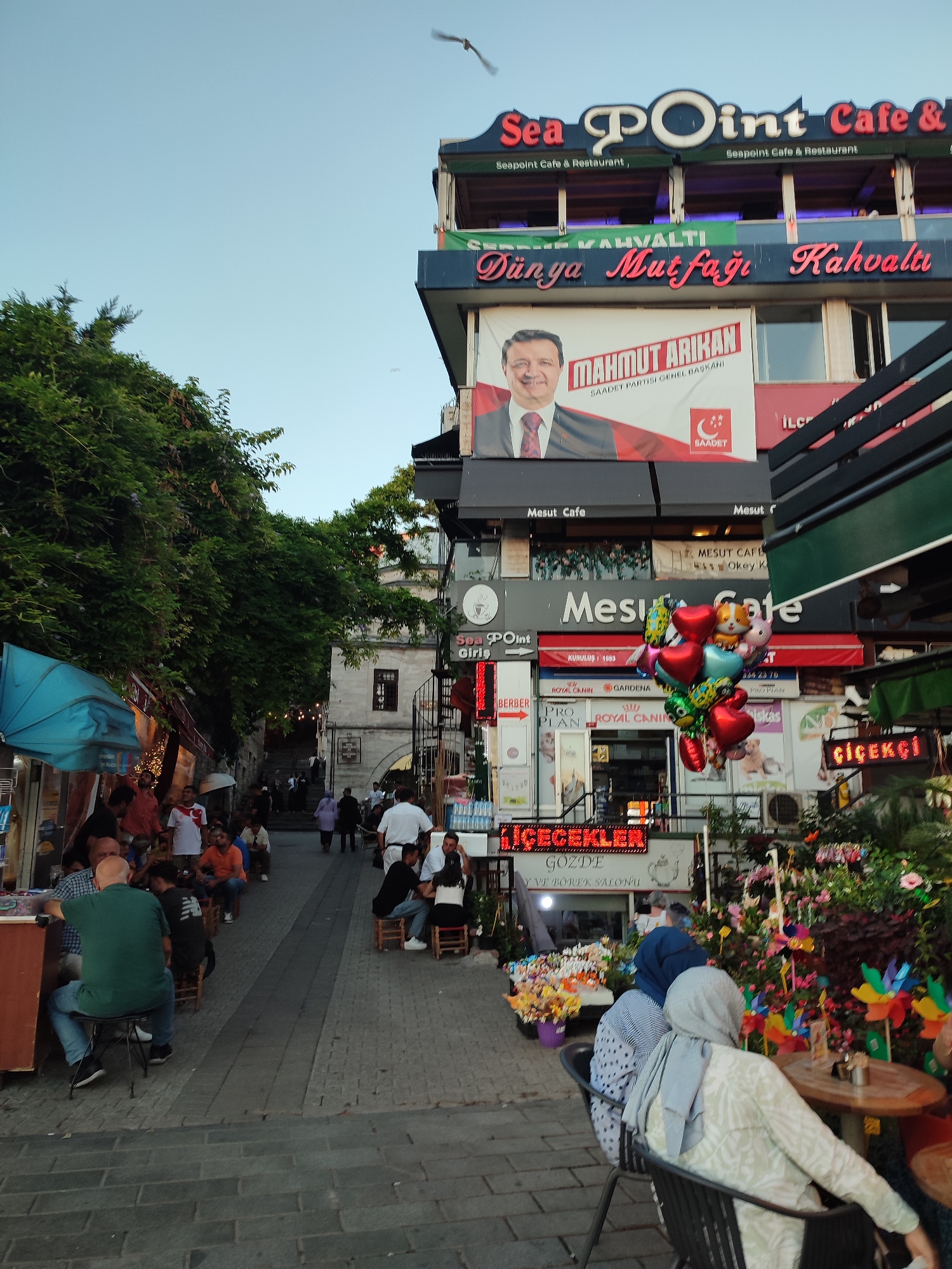 The image depicts a street scene in Üsküdar, Turkey, on a moderately clear day. In the foreground, several people are seated at small tables and chairs on a cobblestone walkway. Some are facing forward, engaged in conversation or observing their surroundings, while others are partially obscured. A man in a green shirt is seated at a stall on the left.
The background is dominated by multi-story buildings with various signs and advertisements. One prominent sign reads "Sea Point Cafe & Restaurant". Below this, another sign features a portrait of a man with the name "MAHMUT ARIKAN" and "SAADET PARTISI GENEL BAŞKANI". Further down, several shops are visible, including one with balloons suspended in front and another displaying flowers and decorative windmills. The architecture suggests a commercial area with a blend of modern and older structures. Lush green trees line the left side of the street, providing shade. The overall atmosphere appears to be that of a bustling urban environment, likely in the evening or late afternoon given the lighting.