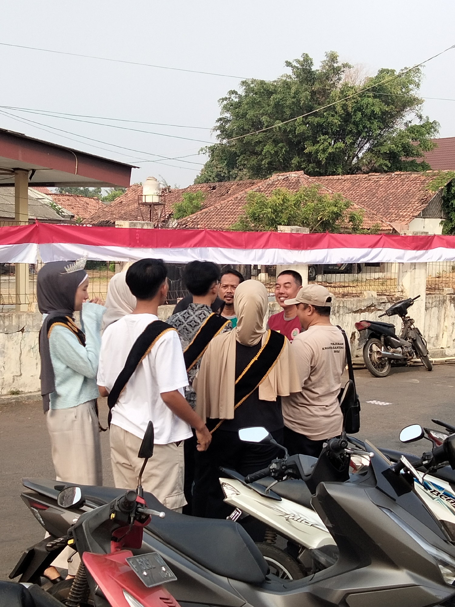 This image shows a group of six people gathered outdoors, possibly for an event, with a red and white banner in the background. The people appear to be conversing. A motorcycle is parked to the right. The location is identified as Cariu, Indonesia. The environment suggests a warm, possibly humid day, with buildings and trees visible. The people are dressed casually, with some wearing headscarves. The red and white banner could indicate a national celebration or event. The presence of motorcycles suggests a common mode of transportation in the area.