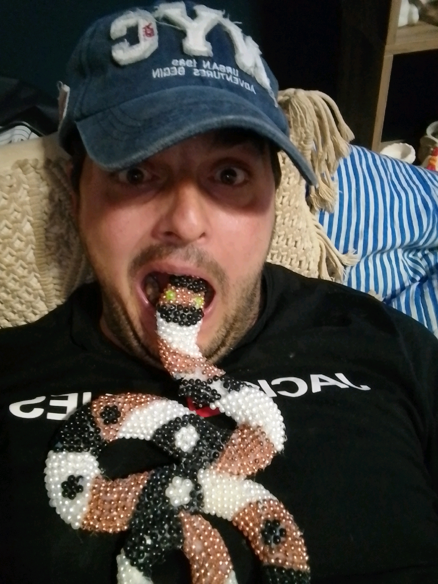 The image shows a man wearing a blue baseball cap with white lettering and a black t-shirt. A beaded snake, with a patterned body of white, black, and copper beads, is positioned so that its head appears to be in the man's mouth. The man's eyes are open, and his mouth is agape around the snake's head. The background is blurred, showing parts of a beige knitted throw and a blue and white striped fabric. There are no discernible elements in the image that would allow for identification of the city of Wedel, Germany.