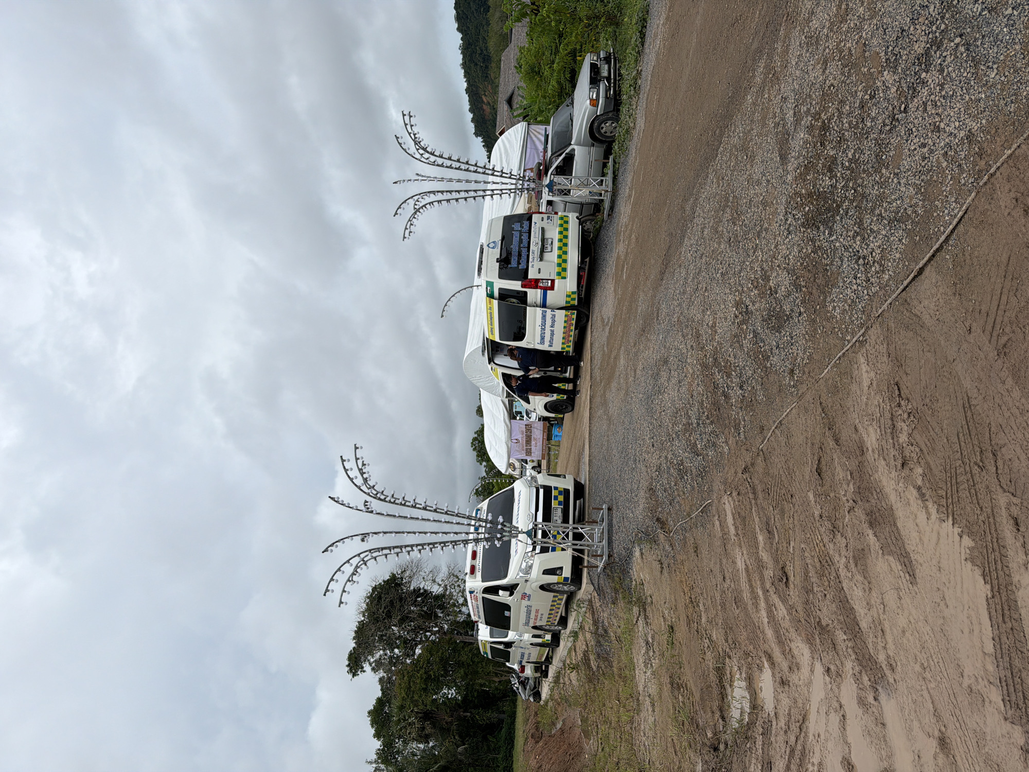 In Si Sunthon, Thailand, on a cloudy day, a scene unfolds involving medical response vehicles and infrastructure. The primary focus is on a convoy of white ambulances, some with red and blue lights illuminated, parked on a gravel road. One ambulance prominently displays the text "Wattanspat Hospital P." and a logo. Adjacent to the ambulances are large white tents, suggesting a temporary medical facility or command center.
In the foreground, a metal structure resembling a decorative light pole with numerous small bulbs is positioned. A silver car is parked near the tents, partially obscured by foliage. Several individuals, some wearing uniforms, are visible near the vehicles and tents, indicating activity related to the medical operation. The environment consists of a rough, unpaved road with muddy patches and tire tracks, bordered by dry grass and trees. In the background, hills and more trees can be seen under the overcast sky. The scene suggests a coordinated effort, possibly for a medical event or emergency response, set up in a semi-rural or undeveloped area.
