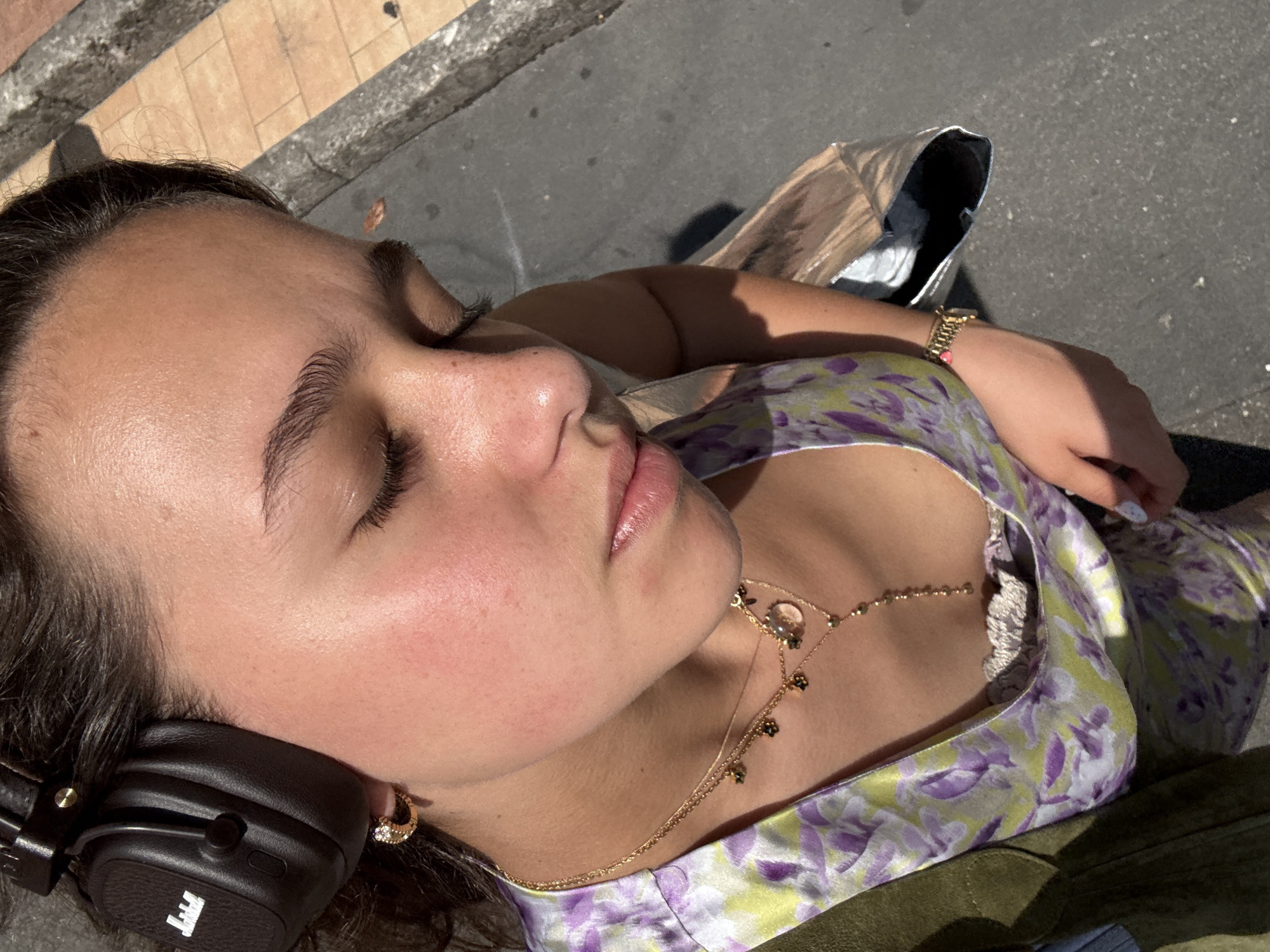 A young female subject is depicted outdoors in what is stated to be Milan, Italy, viewed from an overhead perspective looking downward, with her eyes closed or nearly closed. She wears black Marshall over-ear headphones on her head, with the "Marshall" logo visible on the left earcup. Her dark hair is styled to the sides. A gold hoop earring with a small gem is visible in her left ear. She is wearing a light-colored top with a purple and lime-green floral pattern, layered over a white or cream lace-trimmed undergarment. Multiple gold necklaces are worn, one featuring small dark charms, another with a round, transparent pendant, and a longer Y-shaped chain with small gold beads. A gold watch is present on her left wrist. A portion of an olive-green garment or bag strap is visible on her left shoulder, and a metallic silver-toned bag is seen on the right side of the frame. The background consists of an asphalt surface with a concrete curb and light brown paving stones, illuminated by direct sunlight which casts distinct shadows.