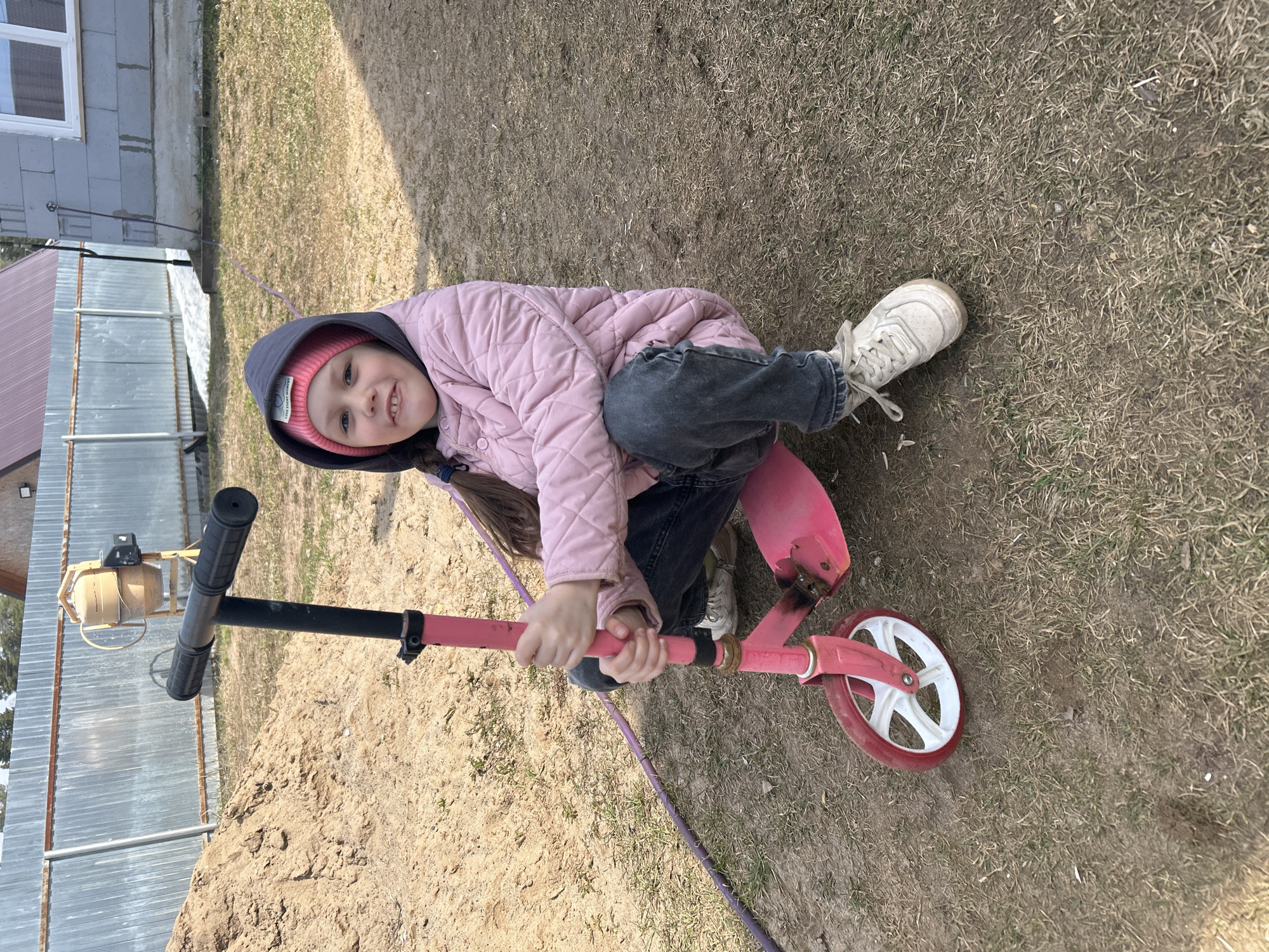 A young girl is depicted outdoors, sitting on a pink and black scooter with white wheels. She wears a pink quilted jacket, a dark hooded sweatshirt, dark pants, and white sneakers. A pink beanie with unreadable white text is visible beneath her hood. She has long brown hair tied in a braid and smiles at the camera, conveying a sense of happiness. Her hands grip the scooter's handlebars.
The setting appears to be a residential or rural area, possibly a yard or an area under development in Chaikovsky, Russia. The ground is a mix of dry, sparse grass and patches of sandy soil. In the background, there's a building with gray cinder block walls and a white window, and a corrugated metal fence. A small, light brown cement mixer or similar construction equipment is visible behind the scooter's handlebars, resting near the fence. A purple hose or cable runs across the ground near the girl. The bright sunlight suggests it is daytime, casting clear shadows.