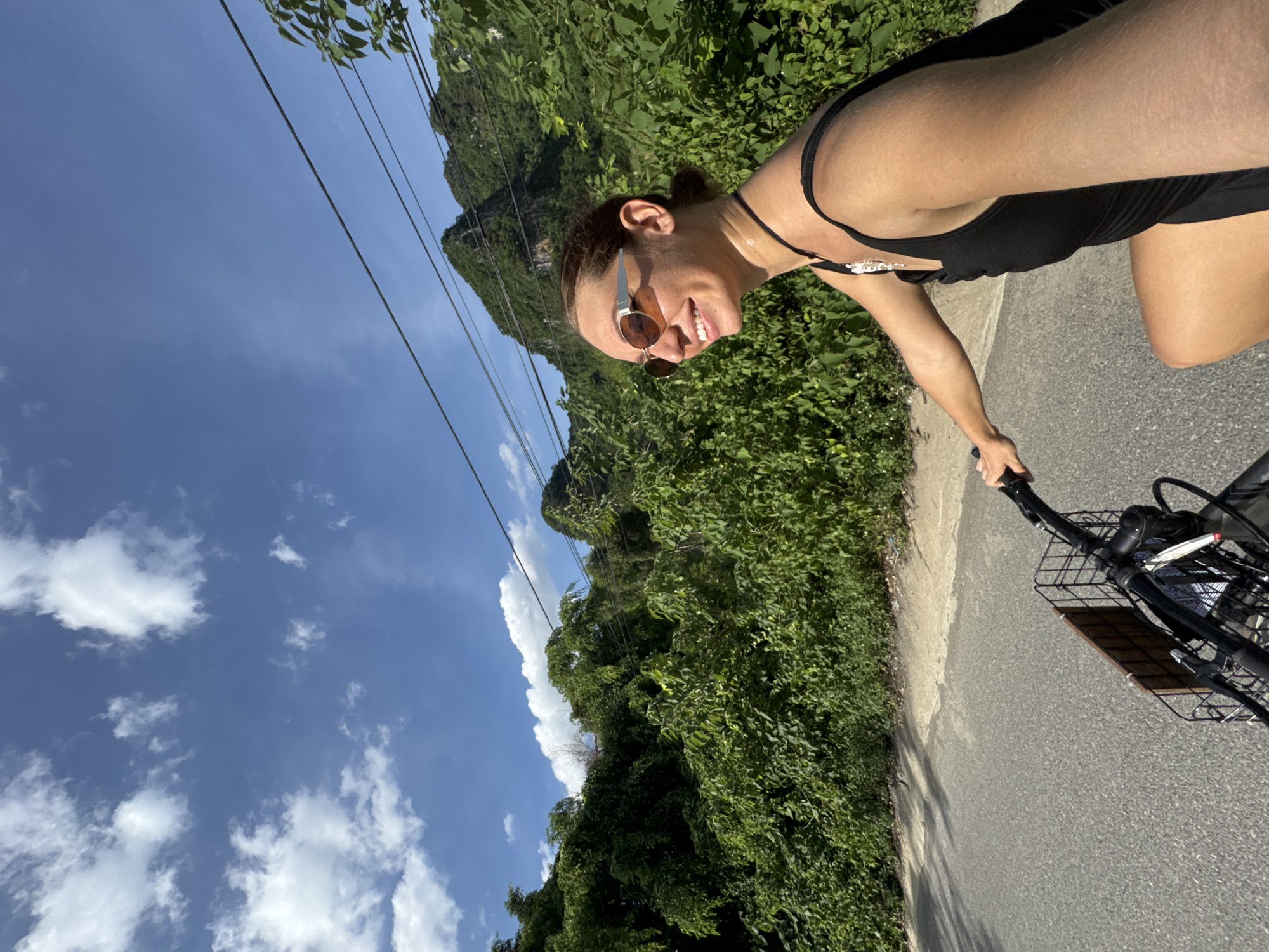 A woman with sunglasses and a black tank top is riding a bicycle on a paved road. She is smiling and looking to her right. The road is lined with lush green trees and vegetation. In the background, there are mountains and a bright blue sky with scattered white clouds. The image appears to be taken in Nam Hoa Lư Ward, Vietnam, based on the context provided, suggesting a rural or mountainous landscape typical of the region. The bicycle has a wire basket attached to the handlebars. The scene depicts a person enjoying an outdoor activity in a scenic natural environment.
