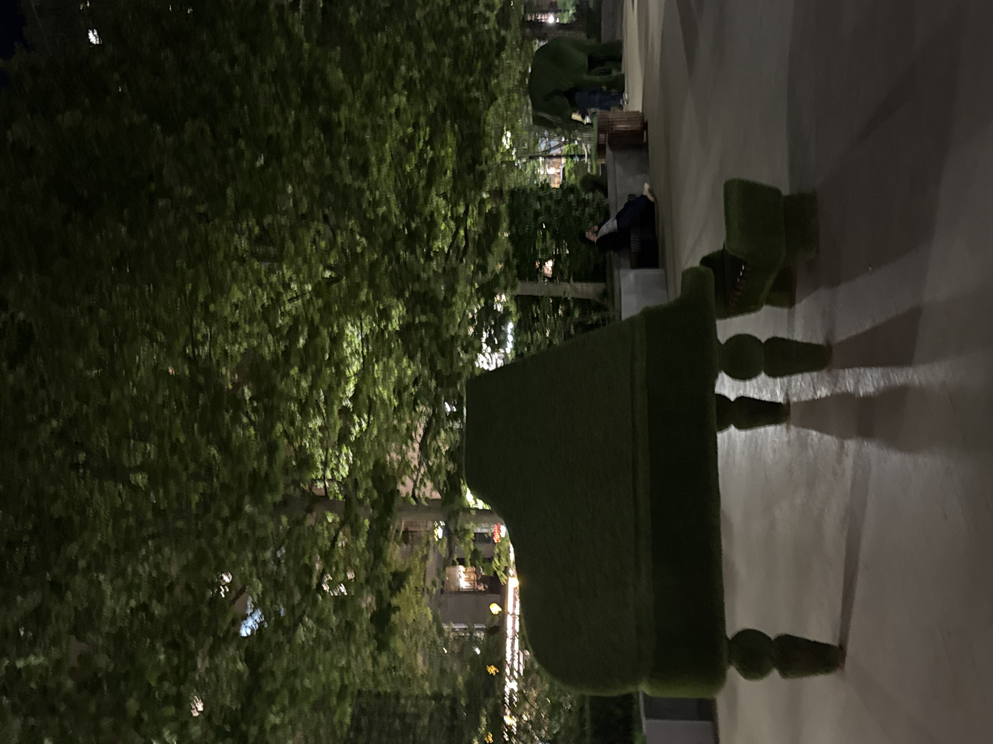 The image shows a dark outdoor scene in Yerevan, Armenia, likely at night. A large, green, grand piano-shaped sculpture covered in artificial turf is in the foreground, casting shadows on the adjacent pavement. A small, matching chair is positioned beside the piano. In the background, a densely foliated tree fills much of the left side of the frame, partially obscuring a building illuminated with lights. People can be seen in the background near the piano, one sitting and another standing near another sculpture.