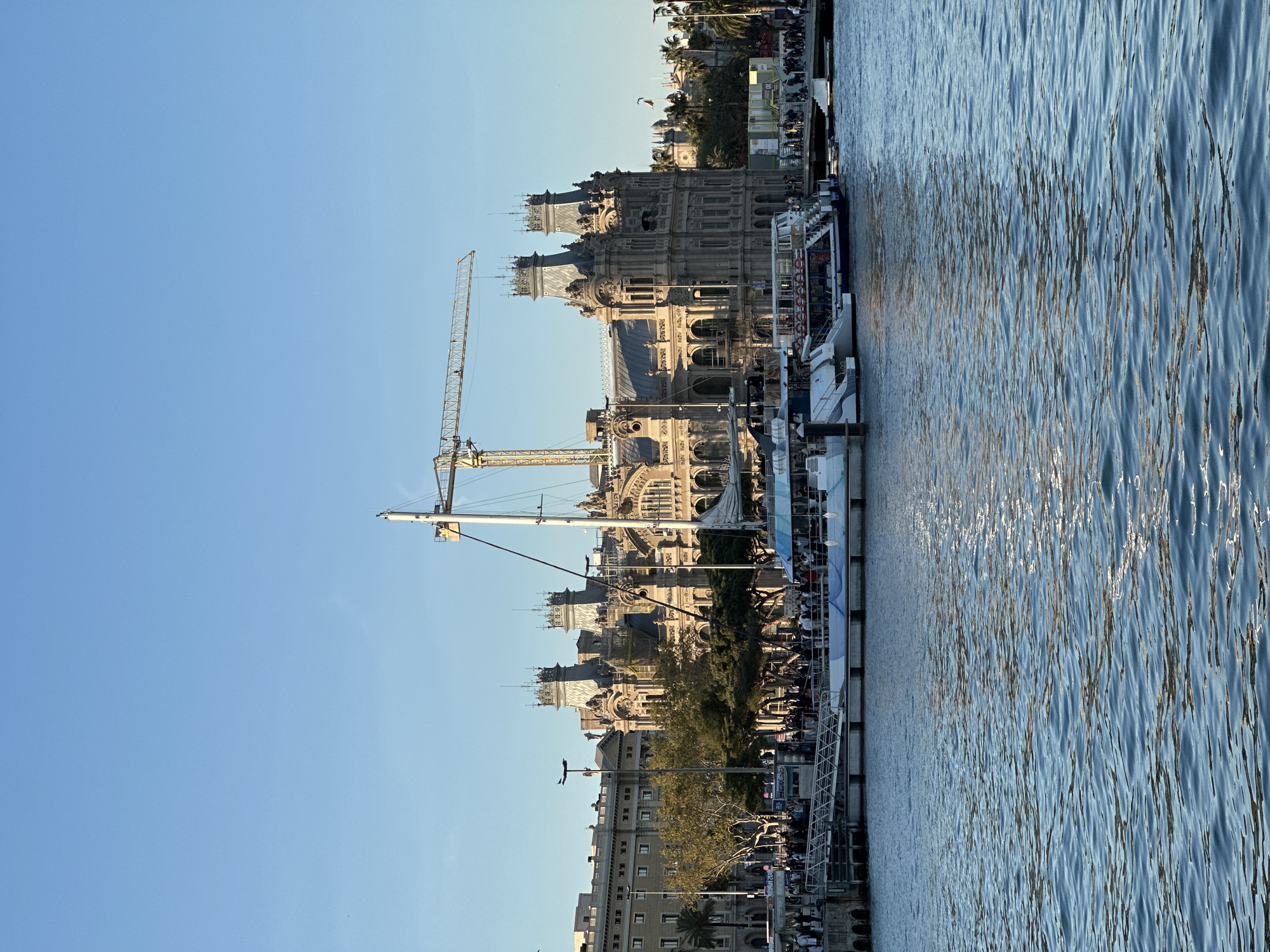 Here is a detailed description of the image:
The image captures a view of Barcelona's waterfront, likely near the Port Vell area. The foreground is dominated by the blue water, displaying rippled reflections. Adjacent to the water is a pier or dock with multiple boats moored alongside it. People are visible along the pier, suggesting a bustling atmosphere.
The backdrop features a historical building with intricate architectural details, potentially the Port Authority building or a similar structure. A large yellow construction crane is also present, indicating ongoing construction or renovation work in the vicinity. Beyond the historical building are other buildings, including some with apartment balconies, that are further back and provide a urban landscape. The sky is a clear, light blue.