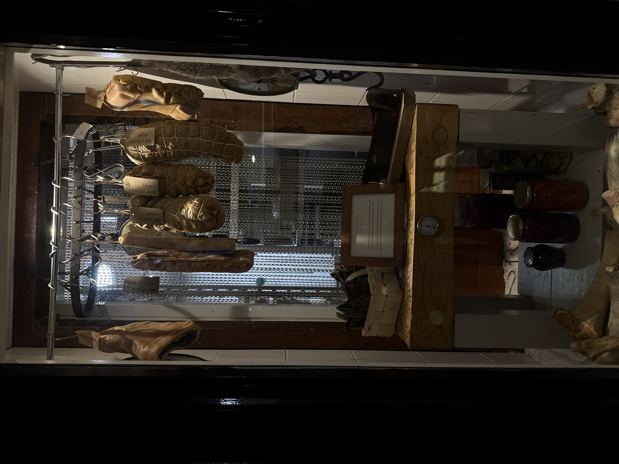 The image depicts a deli display case, likely located in London, United Kingdom, showcasing various preserved meats and pickled goods. On the left, several sausages labeled "GOAT N'DUJA" and "Pancette" are hanging from metal racks. The middle section contains a cleaver, a framed description of "Rugged Hare" emphasizing air-dried beef salums, and a thermometer. Jars containing pickled items are arranged on the right, along with pieces of meat. The display is enclosed in glass and lit dimly, creating a moody atmosphere.