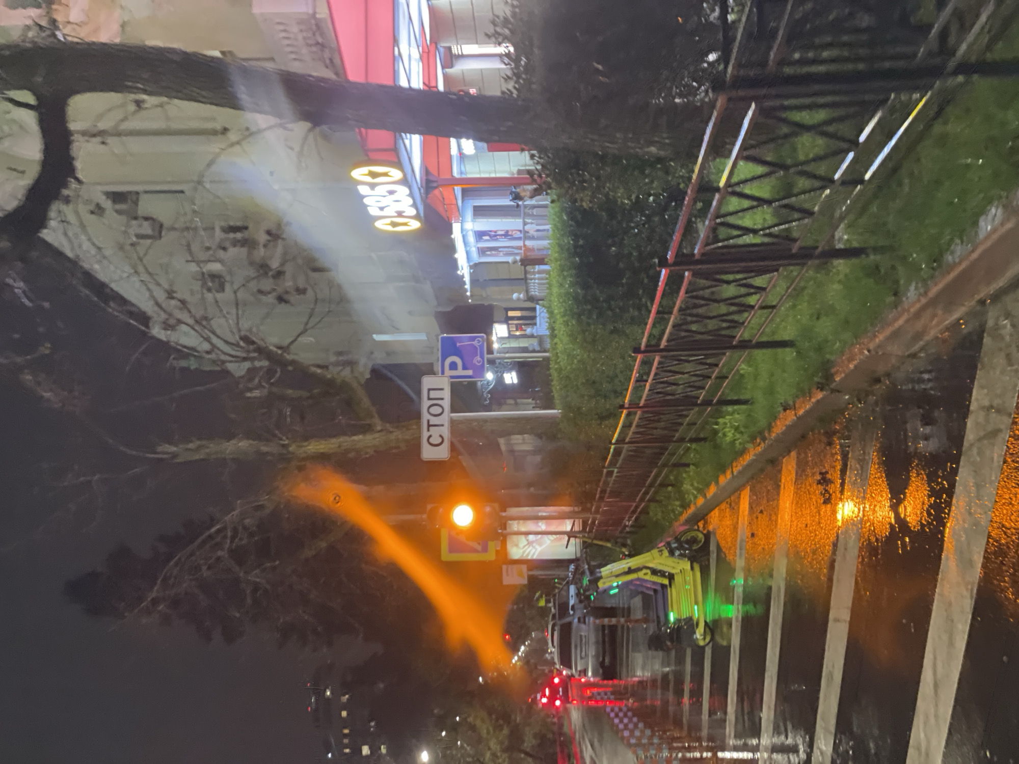 The image shows a street scene at night in Sochi, Russia. It is a wet night, and the street is reflecting the lights. A row of green electric scooters is parked near the curb. A traffic light is visible, displaying an orange light, with a 'СТОП' (STOP) sign nearby. In the background, there is a building with a sign that reads "05850". A metal fence runs along the edge of a grassy area. Trees are on either side of the street. The streetlights and reflections on the wet pavement contribute to the ambiance of the scene. The image is rotated slightly, making the scene appear angled. There is no indication of any people.