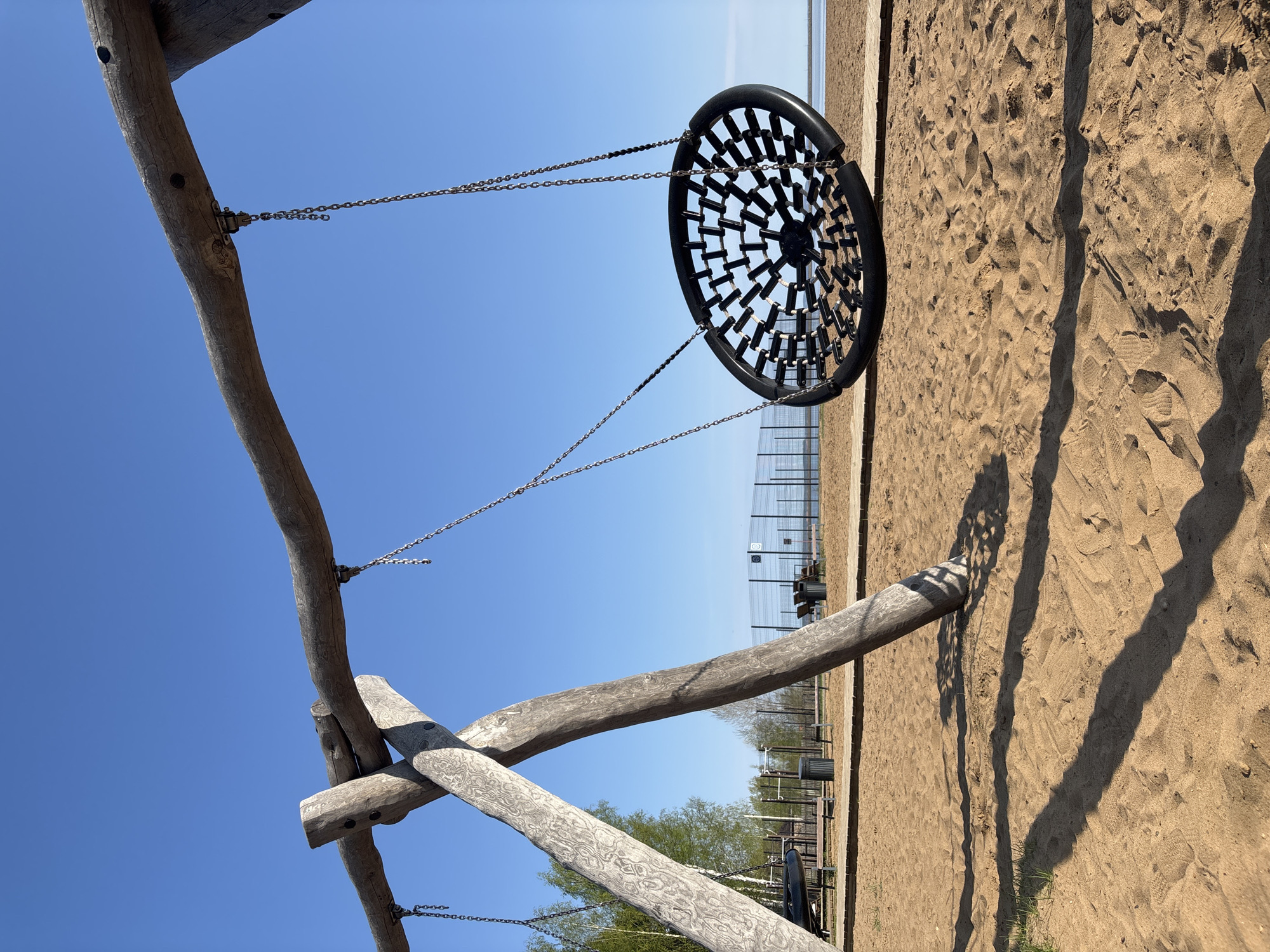 The image depicts a playground in Chaikovsky, Russia. The playground features a wooden structure supporting a large, circular, black swing suspended by chains. The ground is covered in sand. In the background, there is a fence, and behind that, greenery and a body of water can be seen. The sky is clear and blue. There are no people visible in the immediate foreground, but in the background, what may be figures and outdoor furniture are distinguishable. The scene is captured in daylight.