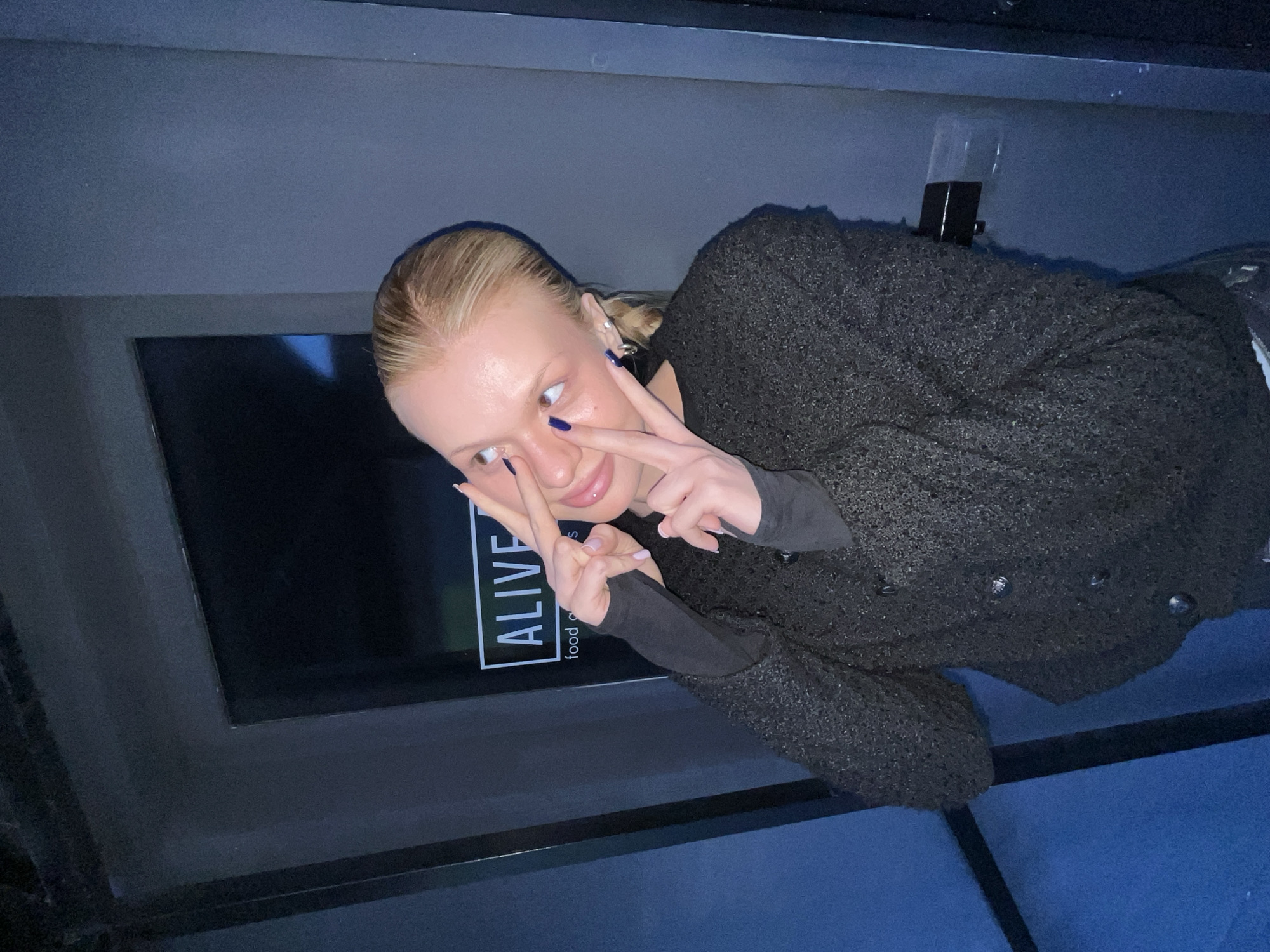 An individual, identified as a young adult female with blonde hair pulled back, is depicted indoors at the ALIVE FOOD establishment located at Lenina Street, 43, Chaikovsky, Perm Krai, Russia. She wears a dark long-sleeved garment underneath a textured, dark grey or black button-up cardigan/jacket. Her fingernails are adorned with dark blue polish. Both of her hands are raised to her face, with the index and middle fingers extended in a "peace sign" gesture, one positioned near her left eye and the other near her right temple. Her eyes are rolled upwards. The background features dark wall panels, and a dimly lit sign displaying "ALIVE" in a white rectangular border, with "food and" visible below, is present to the upper right. A dark door handle is visible on the right side of the frame.