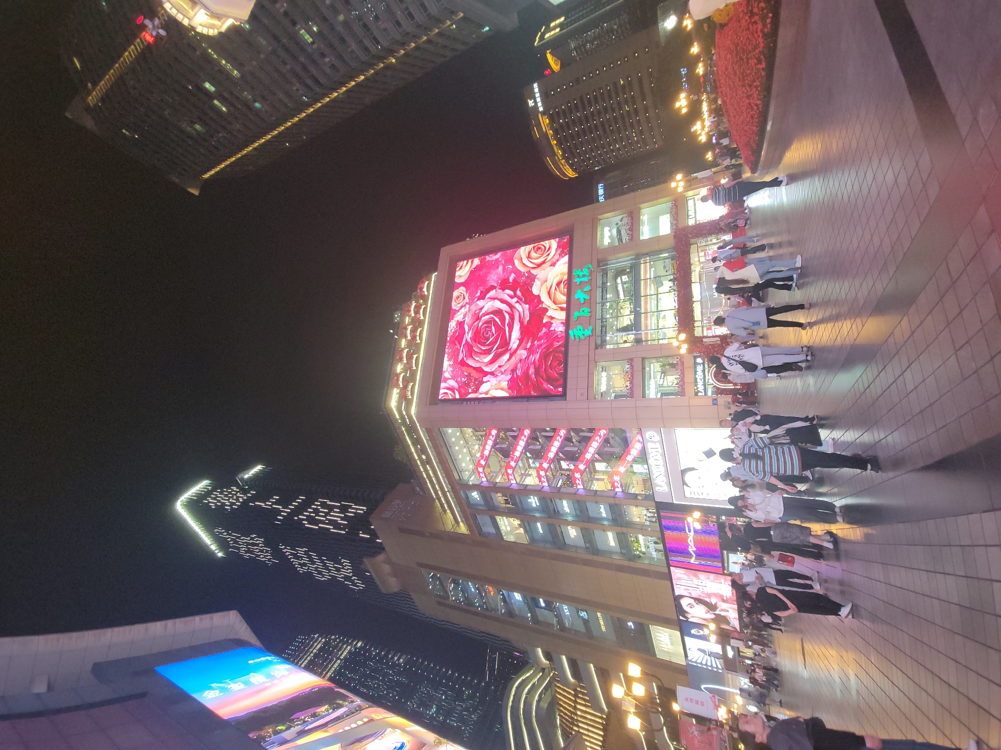 This night-time image captures a bustling urban street scene in Yuzhong District, China. Tall, modern buildings dominate the background, their dark facades punctuated by numerous illuminated windows and decorative lights, suggesting a lively commercial or residential area. The sky is dark, indicating it is late evening or night.
In the mid-ground, a prominent multi-story building features a large, vibrant LED screen on its upper facade, displaying a striking digital artwork of red and pink roses. To the right of this screen, Chinese characters are visible, likely spelling out the name of the establishment, possibly "重庆百货" (Chongqing Department Store). Below this, the building's large glass windows reveal brightly lit interiors, likely retail spaces. Further down the building, the word "LANCOME" can be partially seen on a sign, indicating a luxury brand presence. Another building in the distance also displays illuminated Chinese characters, including "庆银行" (Qing Bank).
A paved pedestrian walkway occupies the right side and foreground, where numerous people are seen walking. They are dressed in casual attire, moving in various directions, suggesting a relaxed evening stroll or shopping activity. Their elongated shadows are cast by the artificial light sources, including streetlights with warm yellow glows and the bright building facades. Red decorative flowers are arranged in planters along the edge of the walkway. The overall atmosphere is vibrant and active, characteristic of a major urban center at night.