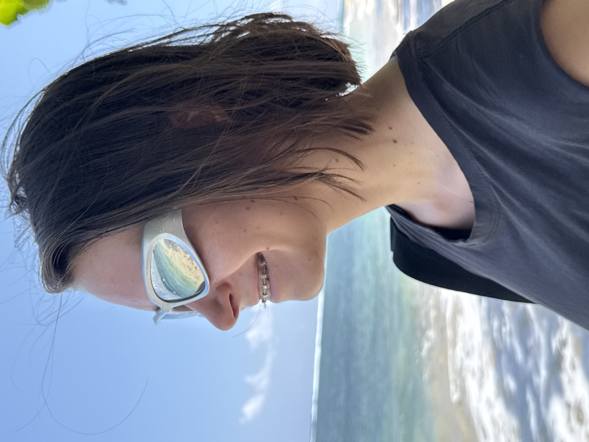 The image, rotated 90 degrees clockwise, presents a close-up profile view of a young individual with dark brown, medium-length hair. The person is wearing light-colored, reflective sunglasses, which distinctly mirror a vibrant blue and green ocean with white breaking waves. A dental brace is visible on their upper teeth, and they are wearing a plain, dark grey or black t-shirt.
The background reveals a sunny coastal environment. A clear, bright blue sky dominates the upper portion, while the lower section features a beautiful ocean with light blue-green waters and white foamy waves along the shore. A small amount of green foliage is visible at the very top of the frame.
The overall scene depicts a relaxing moment at a beach, likely during a sunny day. The reflected ocean in the sunglasses emphasizes the person's engagement with their surroundings. The setting is consistent with a tropical beach, specifically General Luna, Philippines, known for its scenic coastlines. There is no visible text in the image.