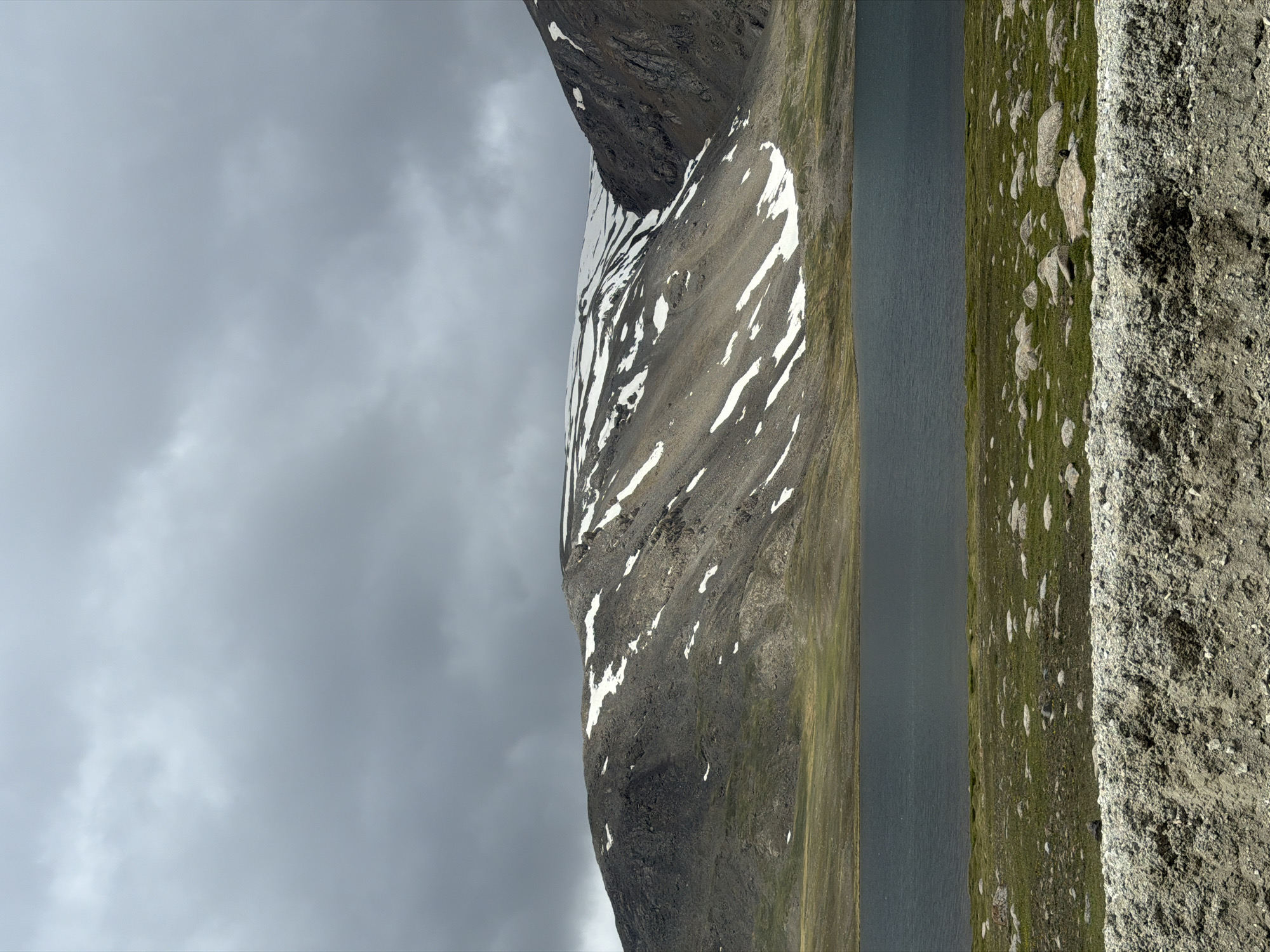 The image depicts a serene mountain landscape with a dark body of water in the foreground, bordered by a grassy bank scattered with rocks. Behind the water, a rugged mountain range rises, its slopes partially covered with patches of snow. The sky above is overcast with thick, grey clouds, suggesting a cooler climate. The foreground also includes a textured, greyish-brown barrier, possibly a retaining wall or natural rock formation. The scene appears to be a natural, undeveloped area, likely in a mountainous region.