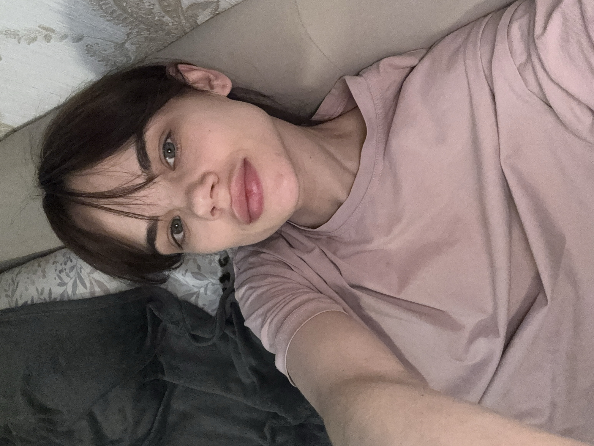 A woman with dark brown hair and green eyes lies on a light gray upholstered surface, possibly a sofa or bed, with a pillow adorned with a floral pattern visible in the upper left corner. She is wearing a light pink T-shirt. A gray blanket is draped over the pillow and a portion of her body. The angle suggests the photo was taken as a selfie. There is no information present to determine the location is Rostov-on-Don, Russia.