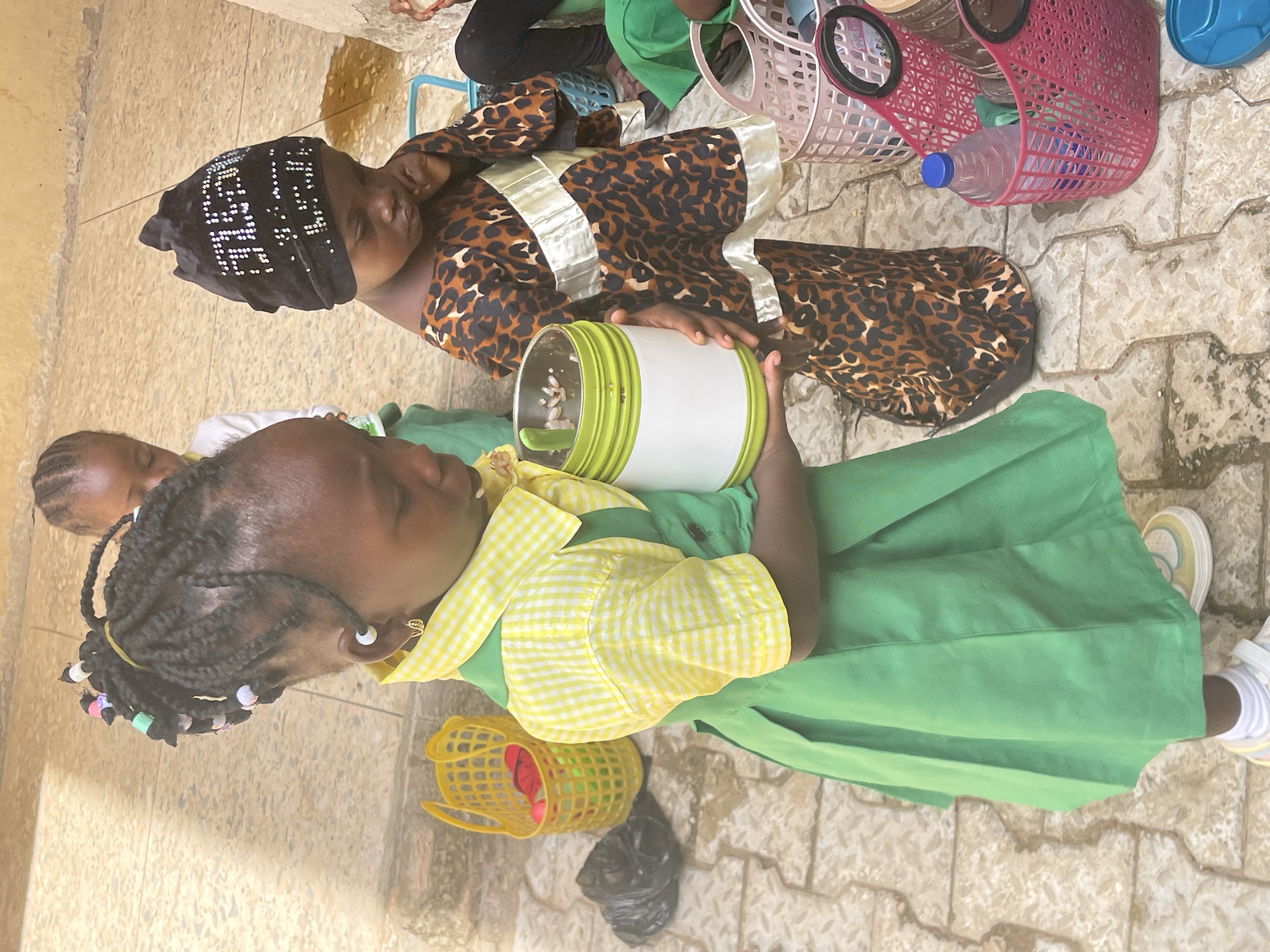 Two young girls are seated on a paved surface in what appears to be a city in Akwanga, Nigeria. One girl wears a green and yellow checkered uniform with her hair in braids adorned with beads. She holds a green and white cylindrical container. The other girl wears a black head covering and a leopard-print dress with a white trim. Nearby are various baskets and a water bottle.