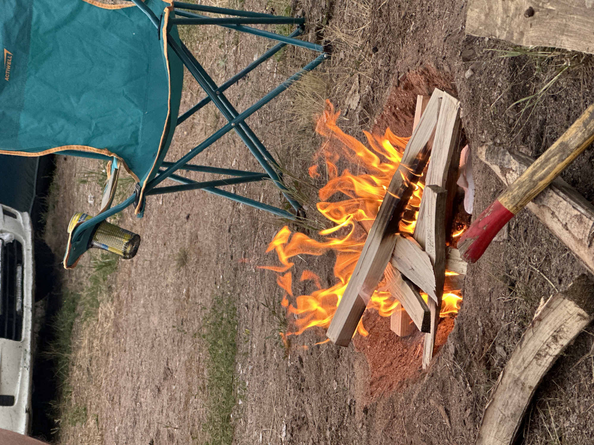 The image shows a campfire in a natural setting, likely outdoors. The fire is burning brightly with flames licking at several pieces of firewood. To the left of the fire is a teal camping chair with orange trim, its legs extending into the dry, brown earth and sparse grass. A white vehicle, possibly a van, is partially visible to the far left.
To the right of the fire, a wooden tool with a red and yellow handle, resembling a shovel or axe, rests on the ground. Additional pieces of wood are scattered around the campfire. The ground appears to be dirt with dry vegetation and some green grass interspersed.
The lighting suggests it might be daytime, possibly late afternoon or early evening, given the warmth of the fire and the light on the objects. The overall scene conveys a sense of outdoor recreation, perhaps camping or a gathering around a fire. There is no visible text in the image except for "ACTIWELL" on the camping chair. The setting appears rustic and possibly remote, consistent with a rural or wilderness location. No people are visible in the image.