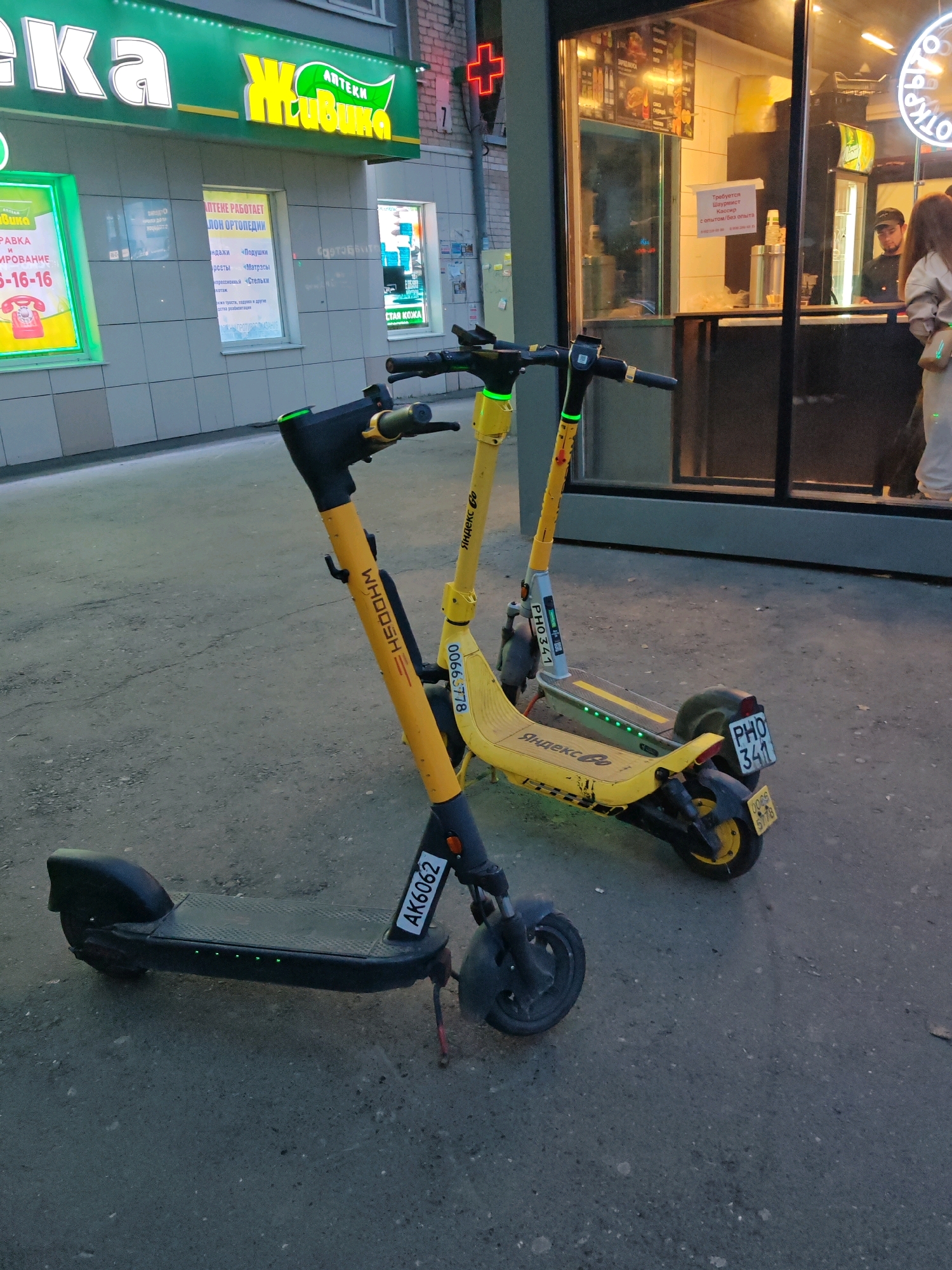 In Yekaterinburg, Russia, two electric scooters, one black and one yellow, are parked on a paved sidewalk. The black scooter, in the foreground, has the license plate "AK6062". The yellow scooter, behind it, has the license plate "PHO 341" and the logo "Yandex Go". Both scooters have green indicator lights on. In the background, a building with green and white signage is visible, with a red neon cross sign on its facade. Adjacent to it is a kiosk with a glass window displaying menus and food items, where a person is visible behind the counter. Another person in a light-colored outfit stands outside the kiosk. The overall scene depicts a typical urban street environment.