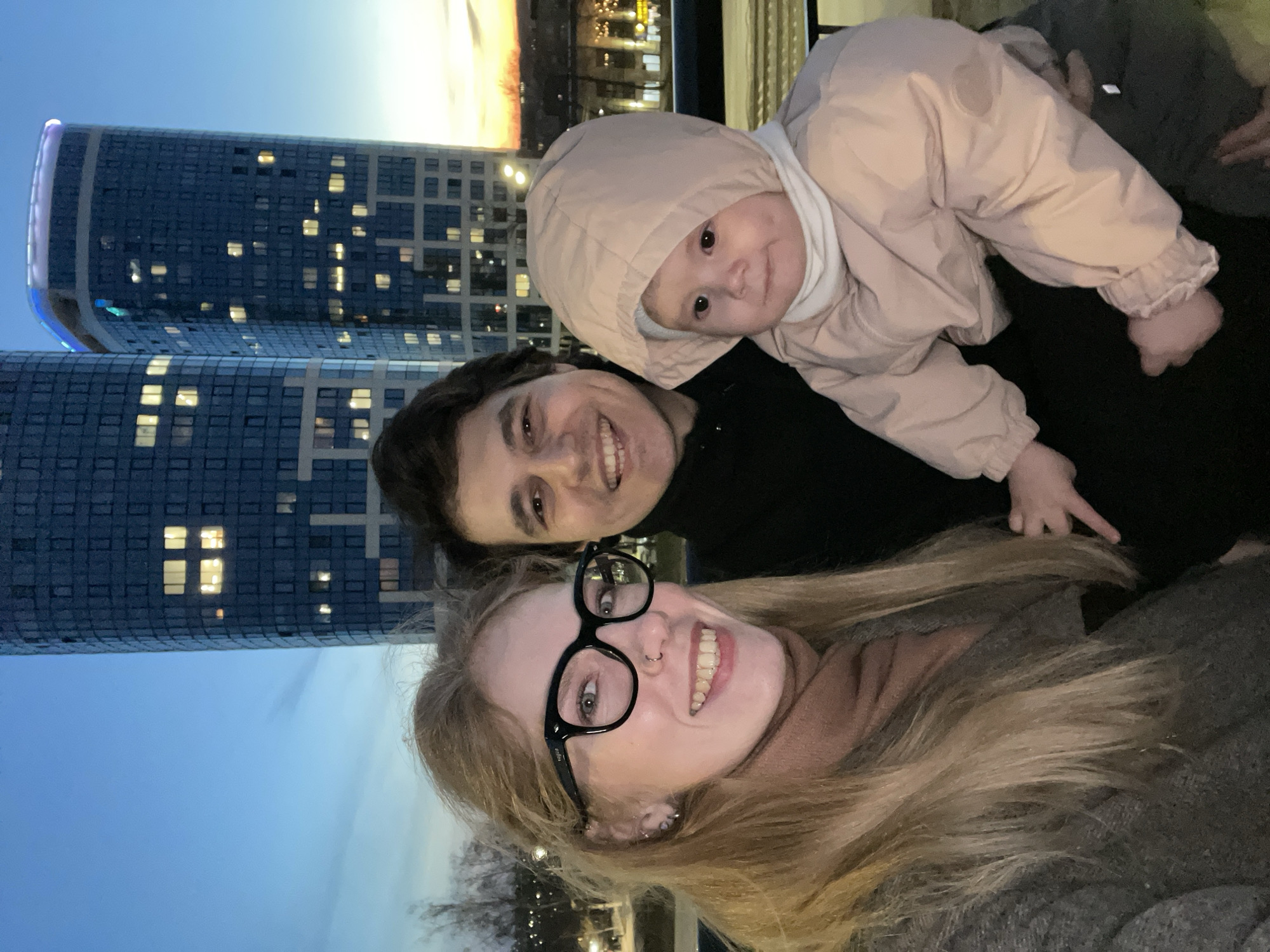 The image is a low-angle selfie taken at night, featuring three people in front of a cityscape. A blonde woman in glasses is in the foreground, smiling. Behind her, a dark-haired person also smiles, holding a baby. The baby, wearing a hooded jacket, looks directly at the camera. The background reveals a tall, modern building with illuminated windows, characteristic of urban architecture. Additional lights can be seen in the distance, suggesting a populated area. Based on visual cues and identifiable landmarks, the location is likely Izhevsk, Russia.