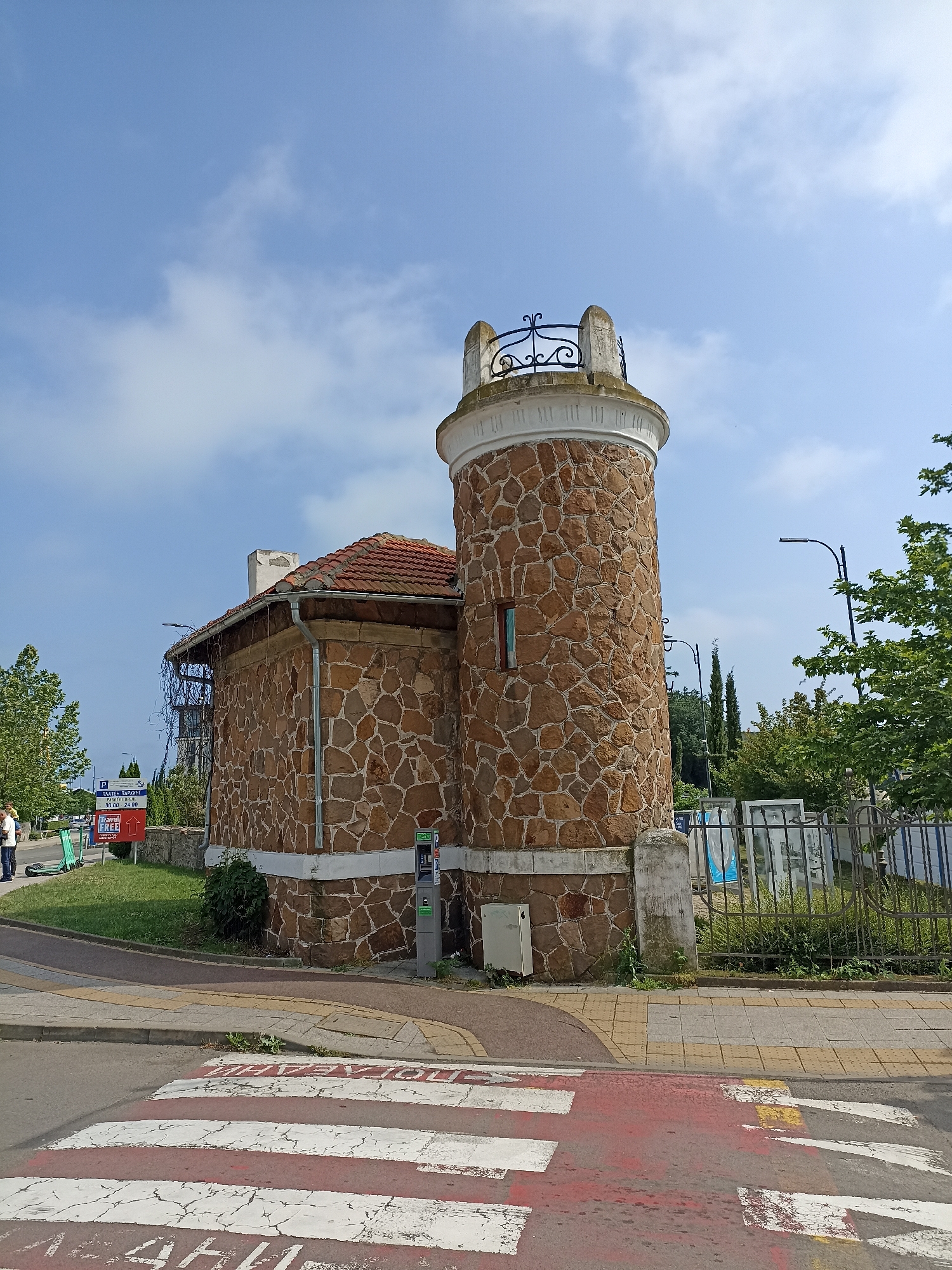 A daytime scene in Burgas, Bulgaria, shows a unique stone-clad building with an attached circular tower. The building's exterior is covered in irregularly shaped, reddish-brown stones, featuring a red tiled roof and a prominent white chimney. The tower is topped with a decorative black wrought-iron railing and has a small, recessed window.
In the foreground, a red and white zebra crossing is painted on the asphalt road, with faded Cyrillic text visible on the red stripes. A paved sidewalk, composed of gray and yellow tiles, runs along the road and curves around the base of the building.
To the left of the building, a grassy area contains a sign written in Cyrillic, stating "ПЛАТЕН ПАРКИНГ" (Paid Parking) with hours "09.00 - 24.00". Below it is a "Travel Free" sign with an upward arrow. Two people are partially visible, along with several green electric scooters on the grass.
To the right of the building, a black metal fence borders a landscaped area with trees, including two tall, slender cypress trees. A modern parking meter or payment kiosk is installed on the sidewalk next to the building's corner, and a white utility box is mounted on the lower section of the tower. Two streetlights are also visible in the background.
The sky is bright blue with scattered white clouds, suggesting clear weather. The image depicts a quiet urban corner, combining historical-style architecture with modern urban infrastructure.