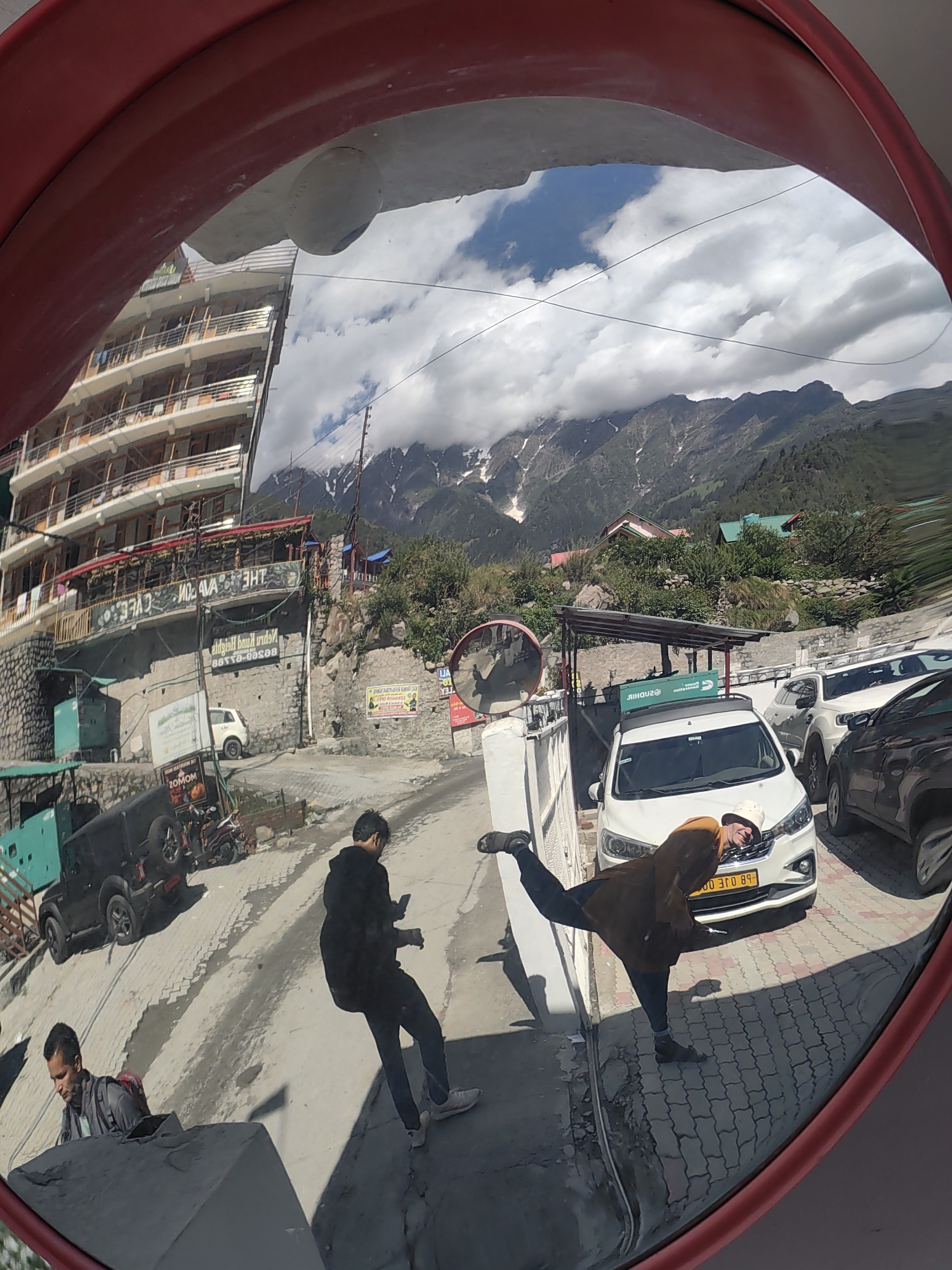 This is a fisheye lens photograph taken in Barheta, India, reflecting in a convex mirror. A multi-story building with balconies is visible on the left, with a sign that reads "THE AVATAR CAFE" in reverse. In the foreground, a man in a black hoodie is captured in mid-stride, looking down at his phone. To his left, another man, wearing a grey backpack, is also looking down, possibly at a phone. Further down the street, a white SUV is parked, and a person in a brown jacket and a bucket hat is performing a dynamic pose, leaning over the hood of the car. The background features a mountainous landscape with snow-capped peaks under a cloudy sky. The road surface is paved with cobblestones.