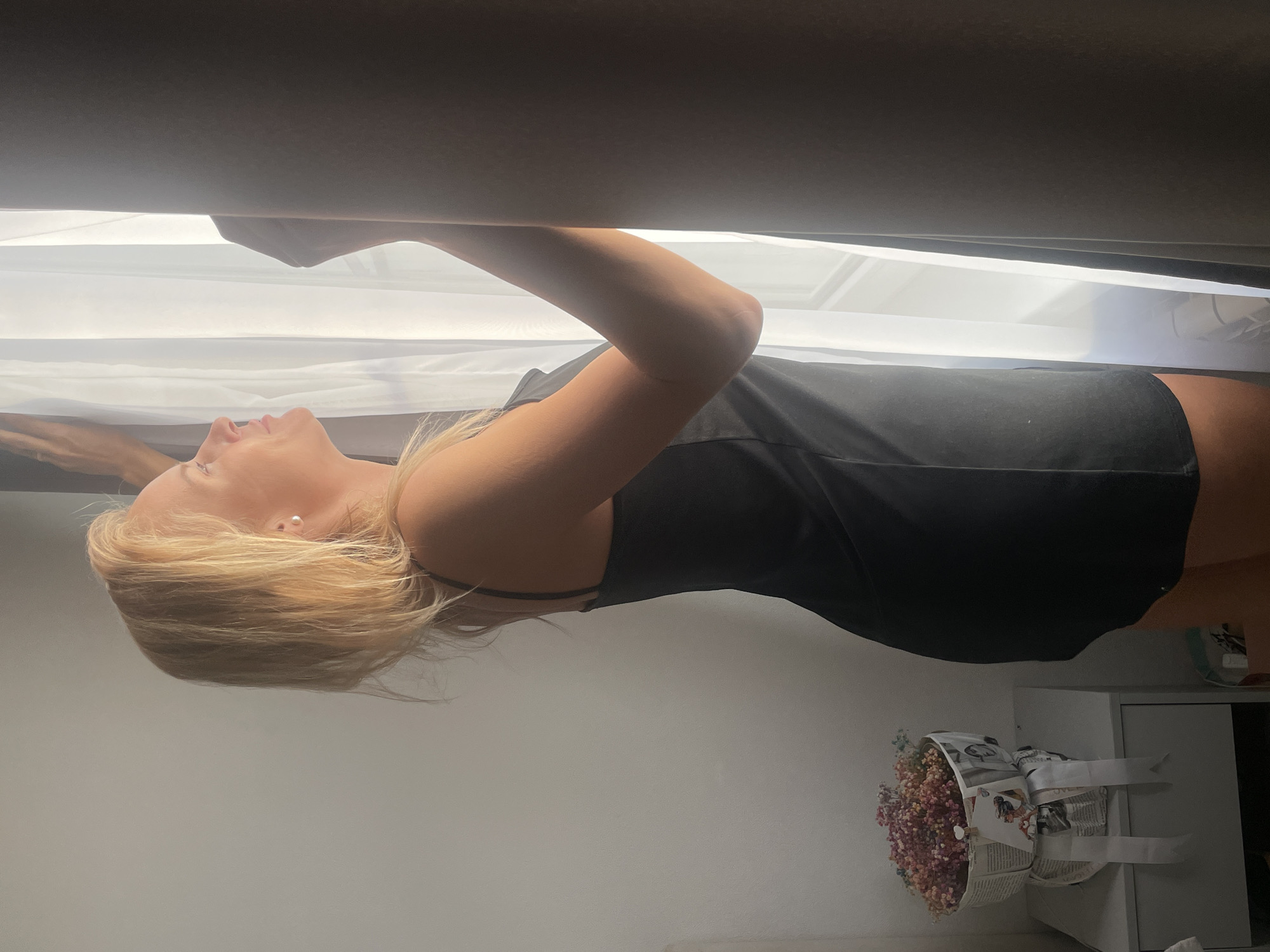 A blonde woman in a black slip dress stands in profile, looking out a window, her hands on the sheer white curtains. Sunlight streams through, illuminating her face and arms. To her left, on a bedside table, is a bouquet of dried flowers wrapped in newspaper and tied with white ribbon. The room appears to be indoors, likely a bedroom, with neutral-colored walls and dark curtains. The location is Baranovka, Russia.