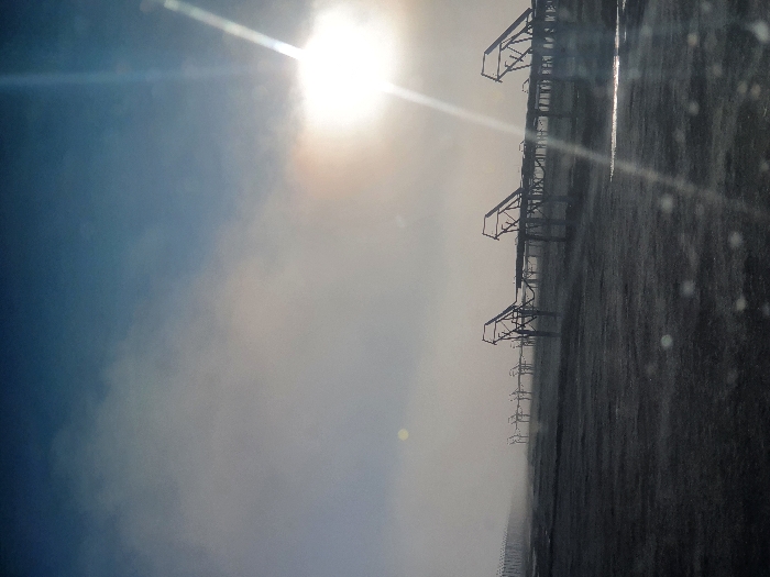A bright sun shines over a desolate, flat landscape. Rows of dark, skeletal structures, resembling industrial equipment, stretch into the distance under a hazy sky. Raindrops or dust particles are visible in the foreground, partially obscuring the view. The overall impression is one of starkness and desolation.