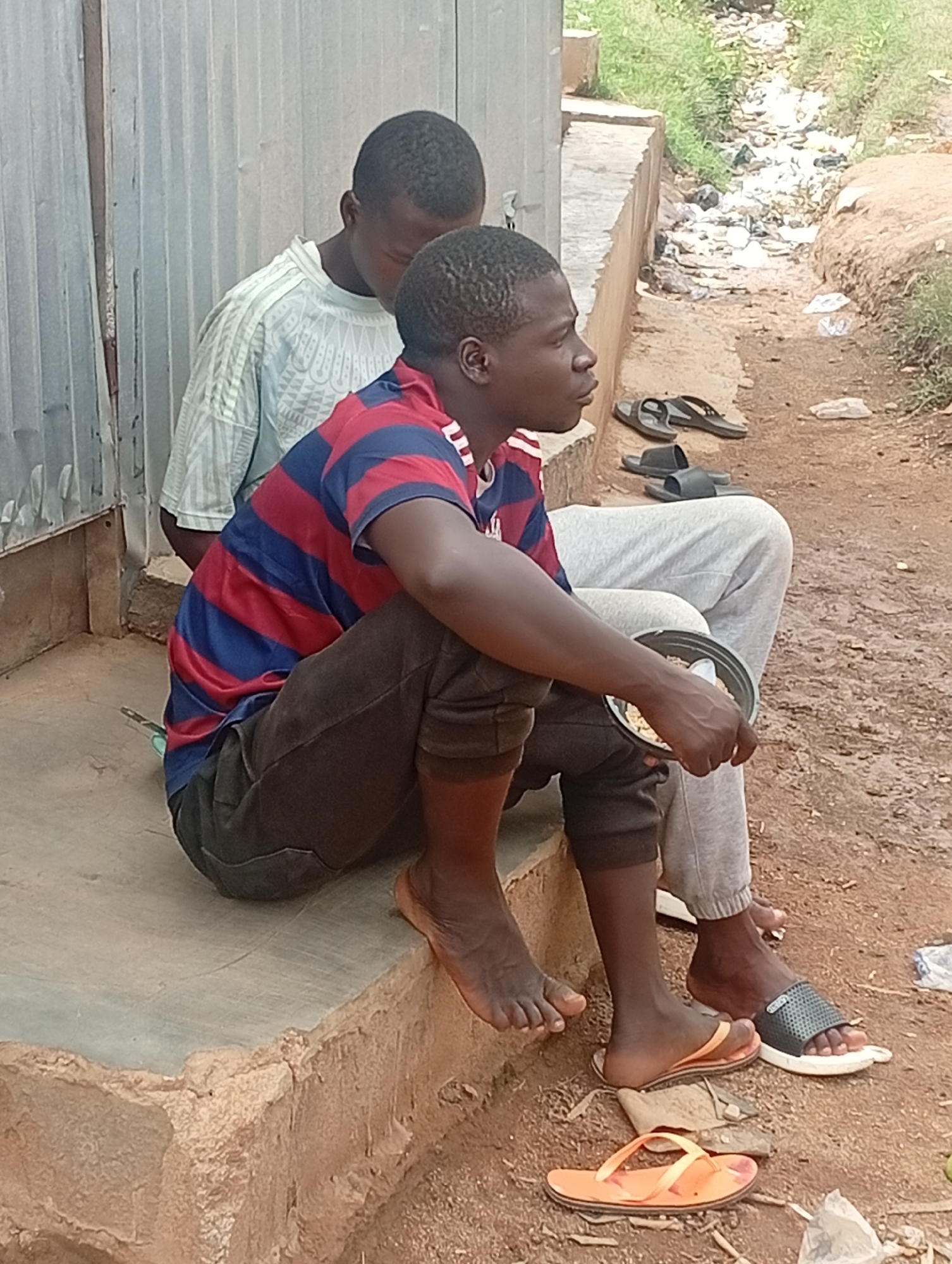 Two young men sit on a concrete ledge in Jos, Nigeria. One wears a striped red and blue shirt and dark pants, holding a bowl and spoon. The other wears a light-colored shirt and gray sweatpants. Several pairs of sandals are scattered on the ground nearby. The setting appears to be a residential area with corrugated metal walls and a ditch. Trash is present in the ditch and on the ground.