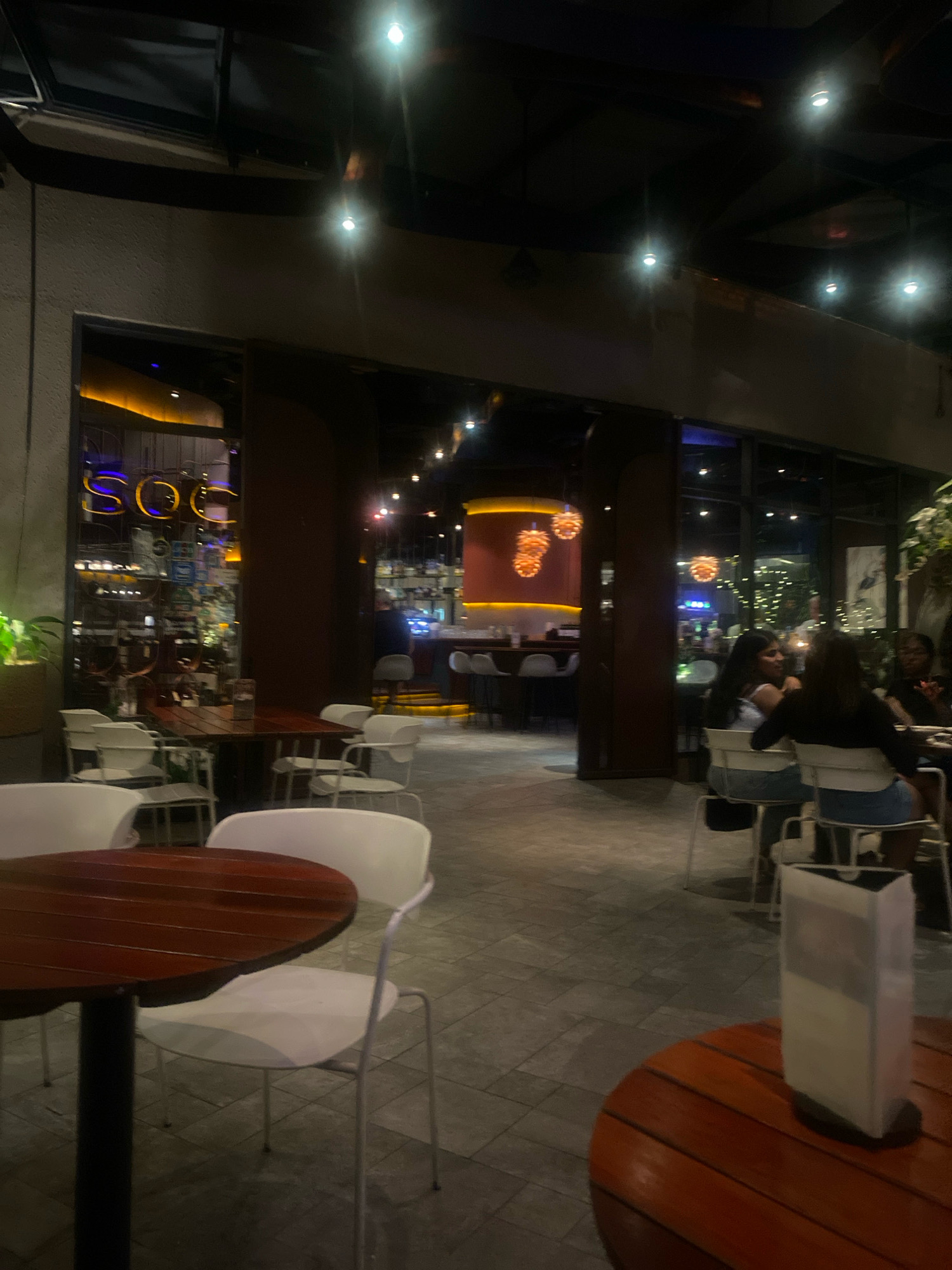 The image depicts an outdoor seating area of a restaurant or bar in Kuala Lumpur, Malaysia, as indicated by the neon signage in the background that reads "SOC". The setting is dimly lit with overhead string lights and accent lighting highlighting architectural features and the bar area. In the foreground, there are several tables and chairs. A round wooden table with a dark central base is in the lower left, with white chairs around it. Another round wooden table with a dark base and slatted top is in the lower right, with a translucent menu holder on it. In the middle ground, there are more white chairs and tables. Through an opening, the interior of the establishment is visible, showing a bar counter with stools and more seating. Several people are seated at tables in the background, with some appearing to be in conversation. The flooring is paved with rectangular tiles. The overall atmosphere suggests a casual evening gathering.