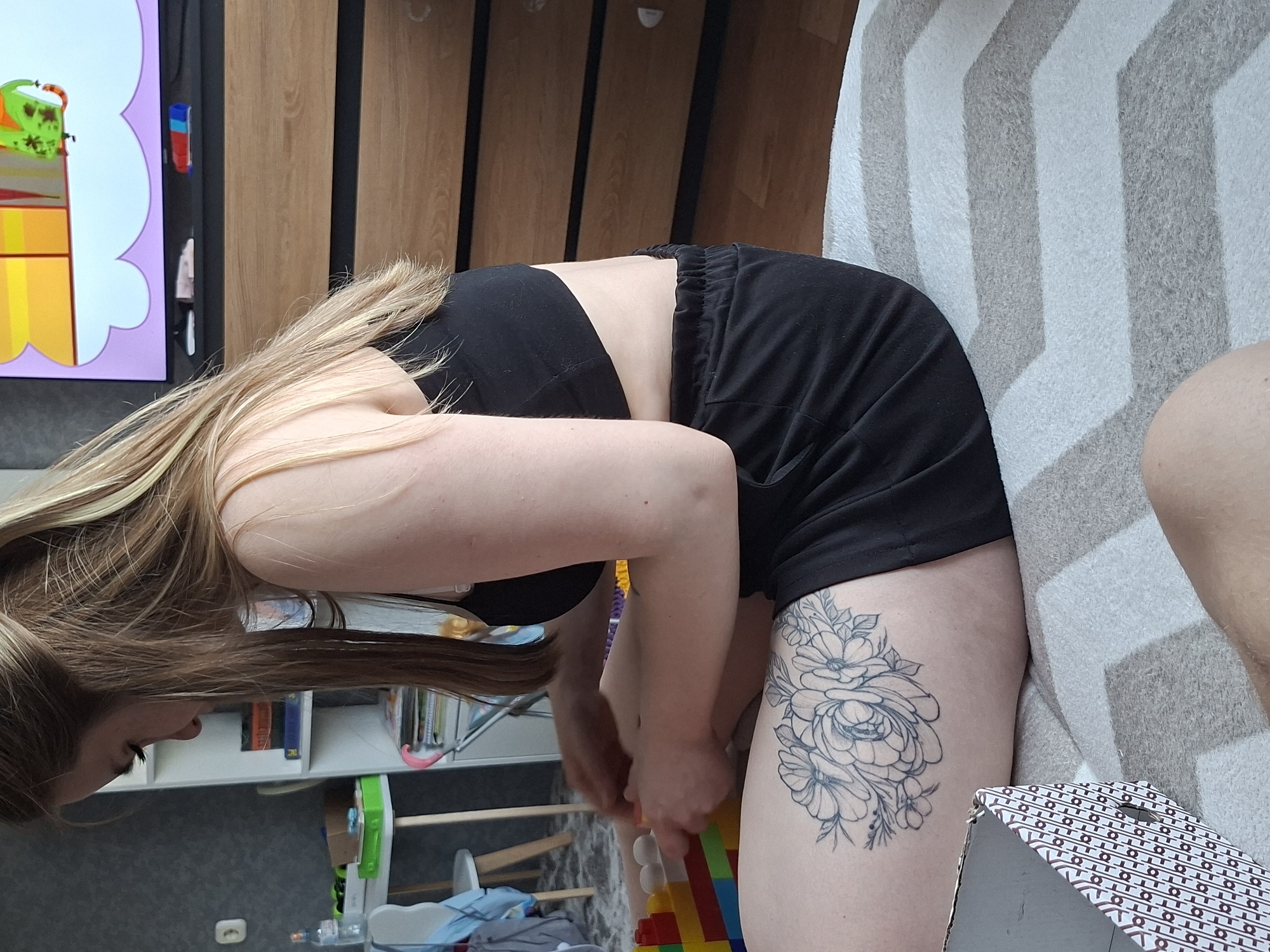 A woman wearing a black top and black shorts is bending over, possibly picking up objects on the floor. She has a floral tattoo on her right thigh. A child's bookshelf is visible in the background, filled with books and toys. In the upper part of the frame is furniture, including a television screen displaying a cartoon. There is also a white and gray striped blanket or sofa next to the woman.
