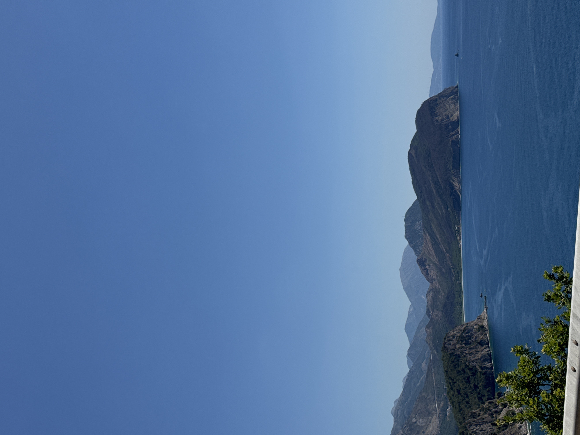 A wide-angle, high-angle shot captures a coastal landscape under a clear blue sky. The scene focuses on the deep blue Adriatic Sea meeting rugged, tree-covered mountains. In the foreground, a white guardrail runs horizontally, with lush green foliage extending into the lower left corner of the frame. A prominent, dark, rocky headland juts out into the sea from the left, its steep cliffs covered in dense vegetation. Beyond this, the coastline curves gently, revealing a sandy beach and more distant, layered mountain ranges fading into the hazy distance. The water is calm, with subtle ripples and a few small, dark rocks visible near the headland. The overall impression is one of a serene, natural coastal environment.