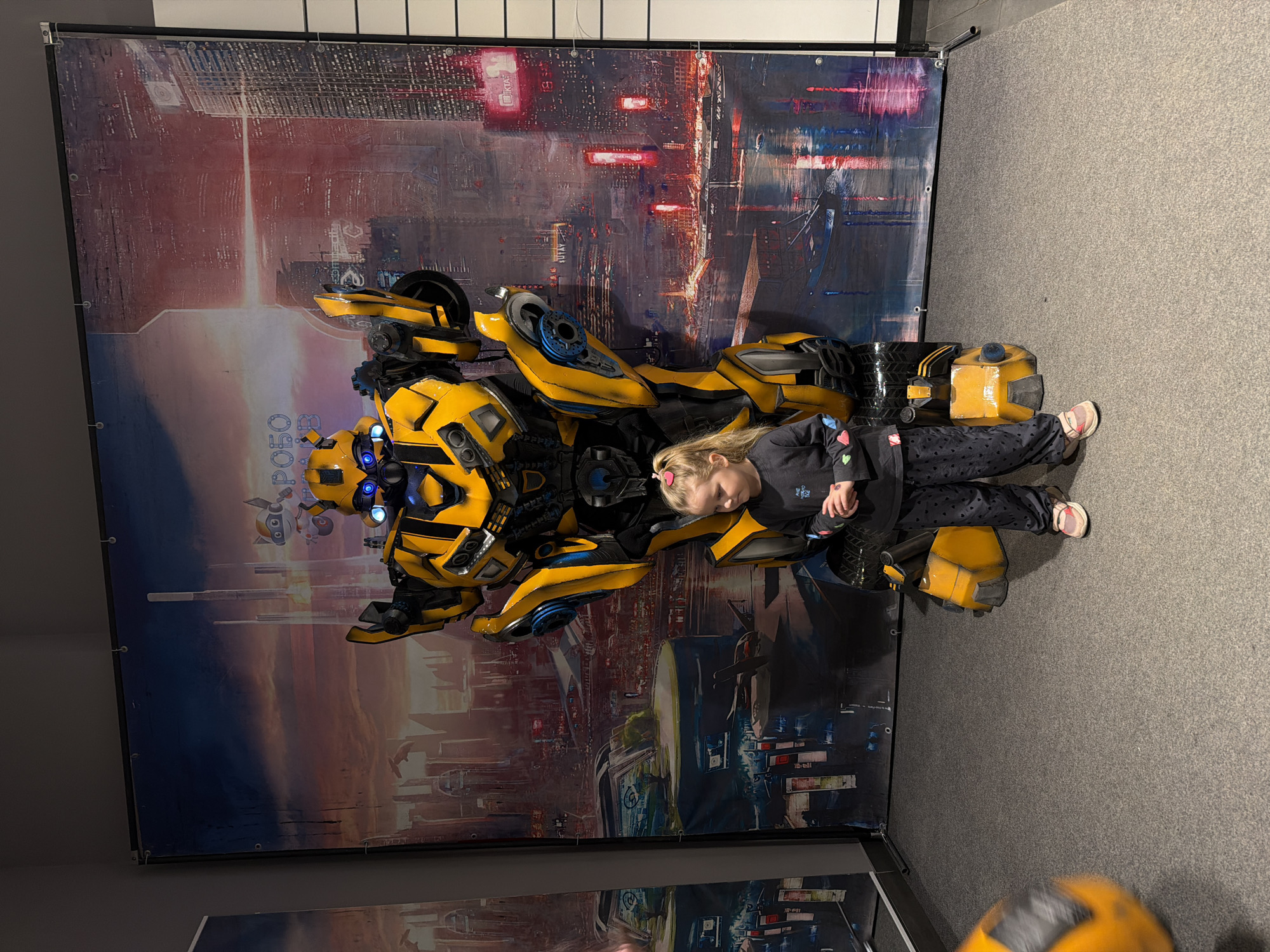 A young girl with blond hair is sitting on a yellow and blue motorcycle-like sculpture in front of a backdrop featuring a futuristic cityscape. She is wearing a dark long-sleeved shirt and patterned pants with light-colored shoes. The backdrop displays an urban scene with tall buildings and what appears to be a sky with a light source emanating upwards. The setting appears to be indoors, likely a display area or exhibition. The floor is carpeted in grey. The location within Kamyshin, Russia is not discernible from the provided image.