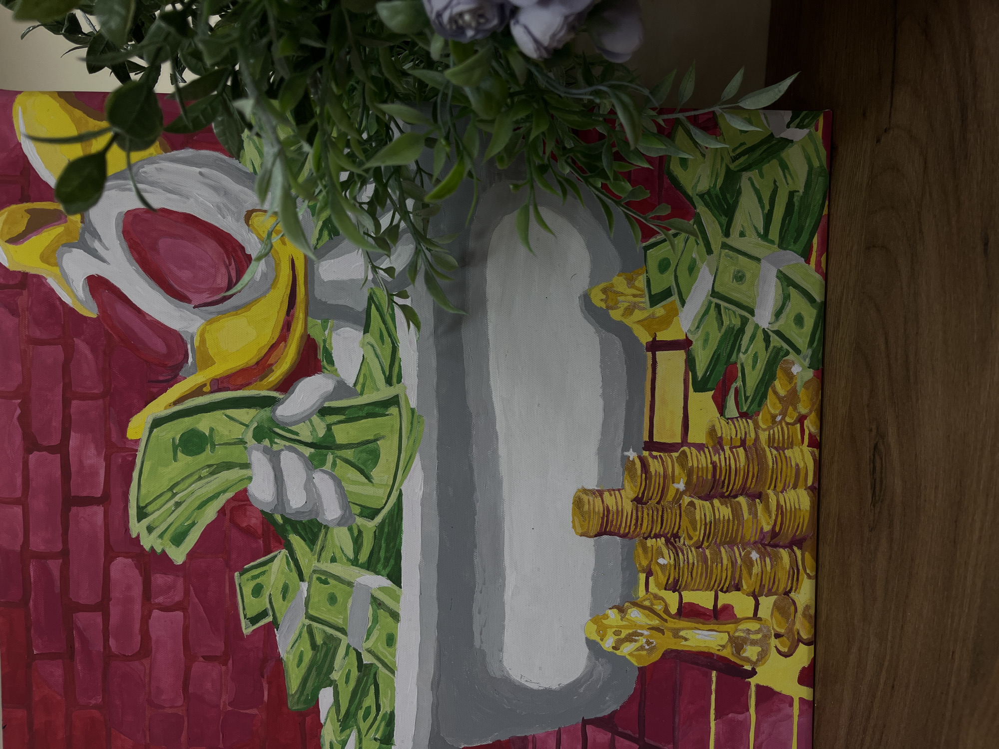 The image shows a painting of a figure with a white and red mask, a grey body, and yellow accents, surrounded by stacks of green money. The painting is displayed indoors, partly obscured by green foliage in the top portion of the frame. The background of the painting is a reddish-purple, stylized brick pattern on the left and a yellow gradient on the right, with gold coins and money. The right side of the frame shows a portion of a wooden surface. There is no information to suggest the location is in the city of Chaikovsky, Russia.