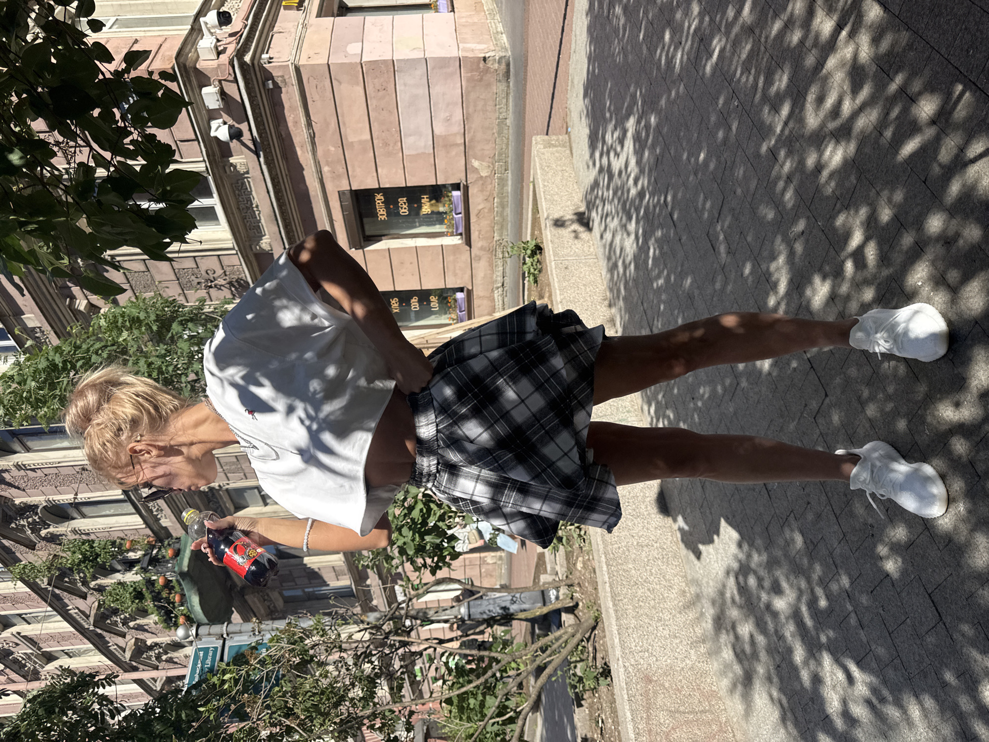 A young Caucasian woman with blonde hair in a messy bun is standing on a paved sidewalk in Saint Petersburg, Russia. She is wearing a white crop top, a black and white plaid skirt, and white sneakers. She is holding a bottle of dark liquid to her lips and looking to her left. The background shows a multi-story building with ornate architecture, trees, and a street sign. The sun is casting shadows on the ground, suggesting it is daytime. The street sign appears to have Russian text. The building has windows and balconies, with plants on some of them. There are security cameras visible on the building. The woman's skin has vitiligo.