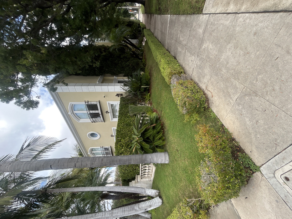 A sidewalk runs alongside a lush green hedge. A two-story building with a beige facade is visible behind the hedge. The building features arched windows, decorative balconies, and ornate trim. Tall palm trees and other foliage surround the building. A large tree provides shade overhead. The scene suggests a residential area with well-maintained landscaping.