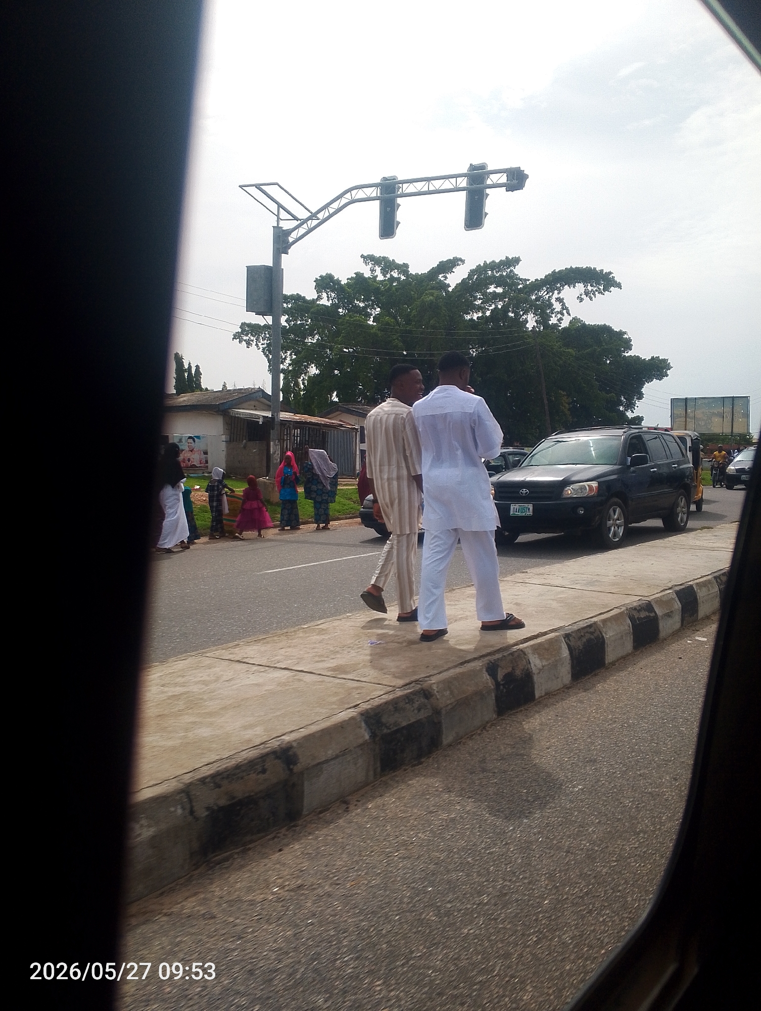 The image depicts a street scene in Lokoja, Nigeria, as seen from inside a vehicle. Two men, dressed in traditional attire, stand on the sidewalk next to a black and white painted curb. One man wears a cream and brown striped outfit, while the other is dressed in white. A group of people, including children and women in headscarves, are walking across the street in the background. A dark-colored SUV is parked on the road, with its license plate partially visible. A traffic light structure is visible overhead. The sky is overcast, and trees are visible in the background. The date and time stamp "2026/05/27 09:53" is at the bottom left corner of the image.