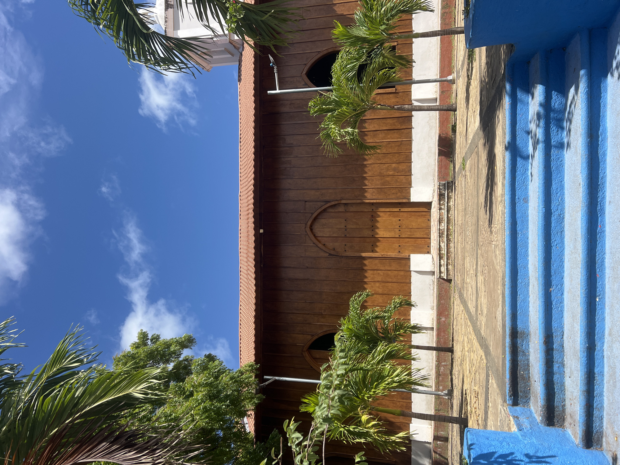 In San Juan del Sur, Nicaragua, a wooden building with a red tiled roof is captured under a clear blue sky with scattered white clouds. The building features large double doors and arched windows, suggesting it might be a church or a similar communal structure. In the foreground, vibrant blue steps lead up to a patterned walkway. Several palm trees are scattered around the building, and foliage from other trees frames the top of the image. The scene is bathed in bright sunlight, casting shadows on the ground.