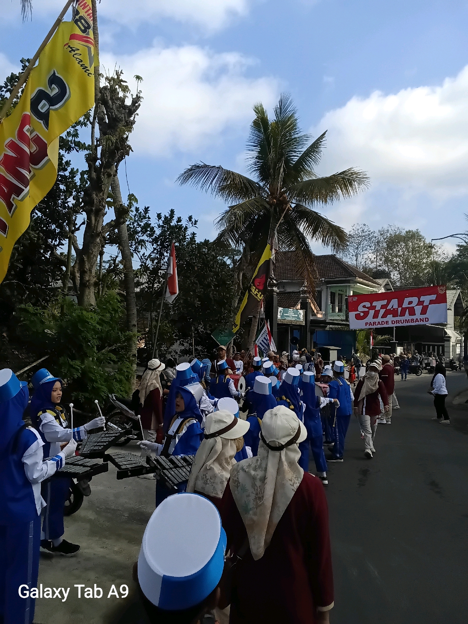 In Bendo, Indonesia, a marching band parade is underway, as evidenced by the "START PARADE DRUMBAND" banner displayed prominently. The parade features individuals, primarily young women, dressed in blue and white uniforms, some wearing hijabs. They are seen marching in formation, some holding what appear to be musical instruments like xylophones and drumsticks. The background shows a street lined with trees, houses, and a coconut tree. The sky is partly cloudy with a bright sun. The overall atmosphere suggests a festive event.