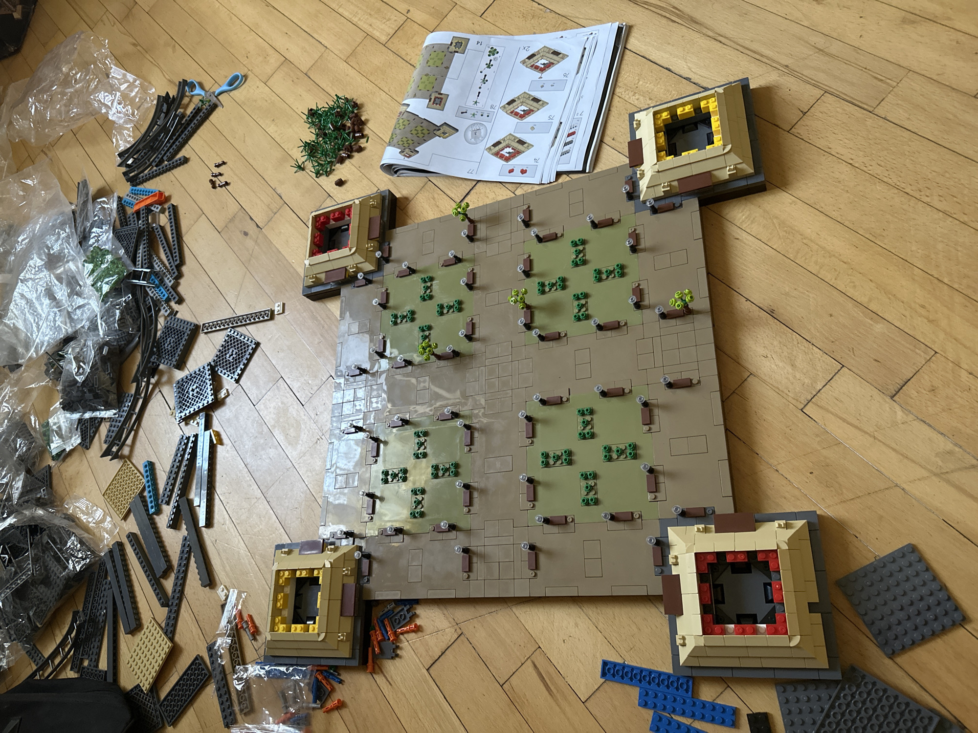 The image shows a partially constructed Lego model on a wooden floor. The model appears to be a city or town layout with several small buildings, some with green foliage representing trees or plants. There are also loose Lego bricks and instructions scattered around the model. The scene is brightly lit, suggesting an indoor environment. There are no people or animals visible in the image. The location in the city of Yerevan, Armenia, cannot be determined from the provided image.