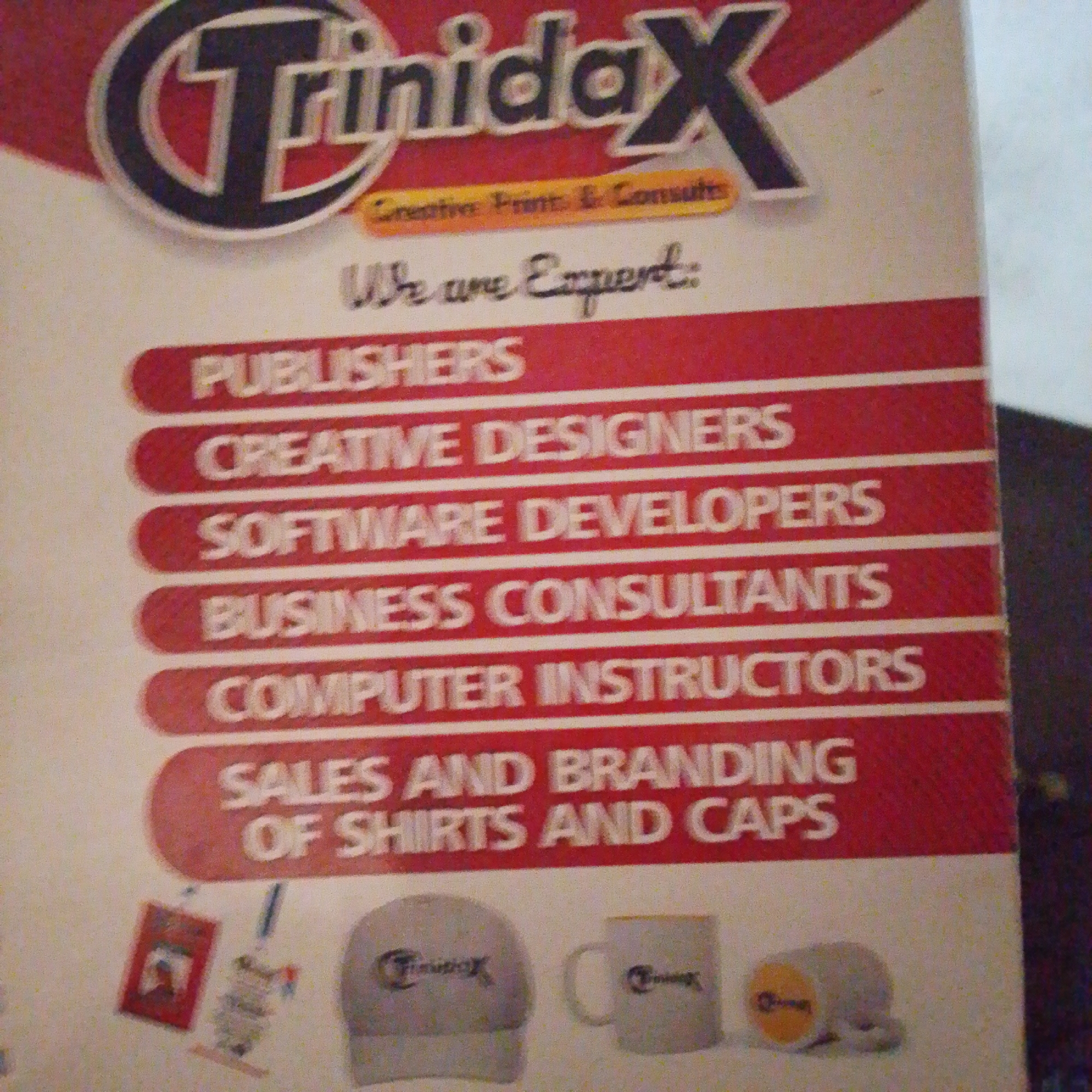 The image displays a colorful flyer for a company named "TrinidadX" which offers "Creative Print & Consults." The flyer lists various services including "PUBLISHERS," "CREATIVE DESIGNERS," "SOFTWARE DEVELOPERS," "BUSINESS CONSULTANTS," "COMPUTER INSTRUCTORS," and "SALES AND BRANDING OF SHIRTS AND CAPS." Below these services, there are images of promotional items such as a cap, a mug, and a plush toy, all bearing the company logo. The background is a light cream color with red and white stripes accentuating the list of services. The flyer itself appears to be a physical item, possibly a printed advertisement or brochure. The context of Jalingo, Nigeria is not visible or derivable from the provided image.