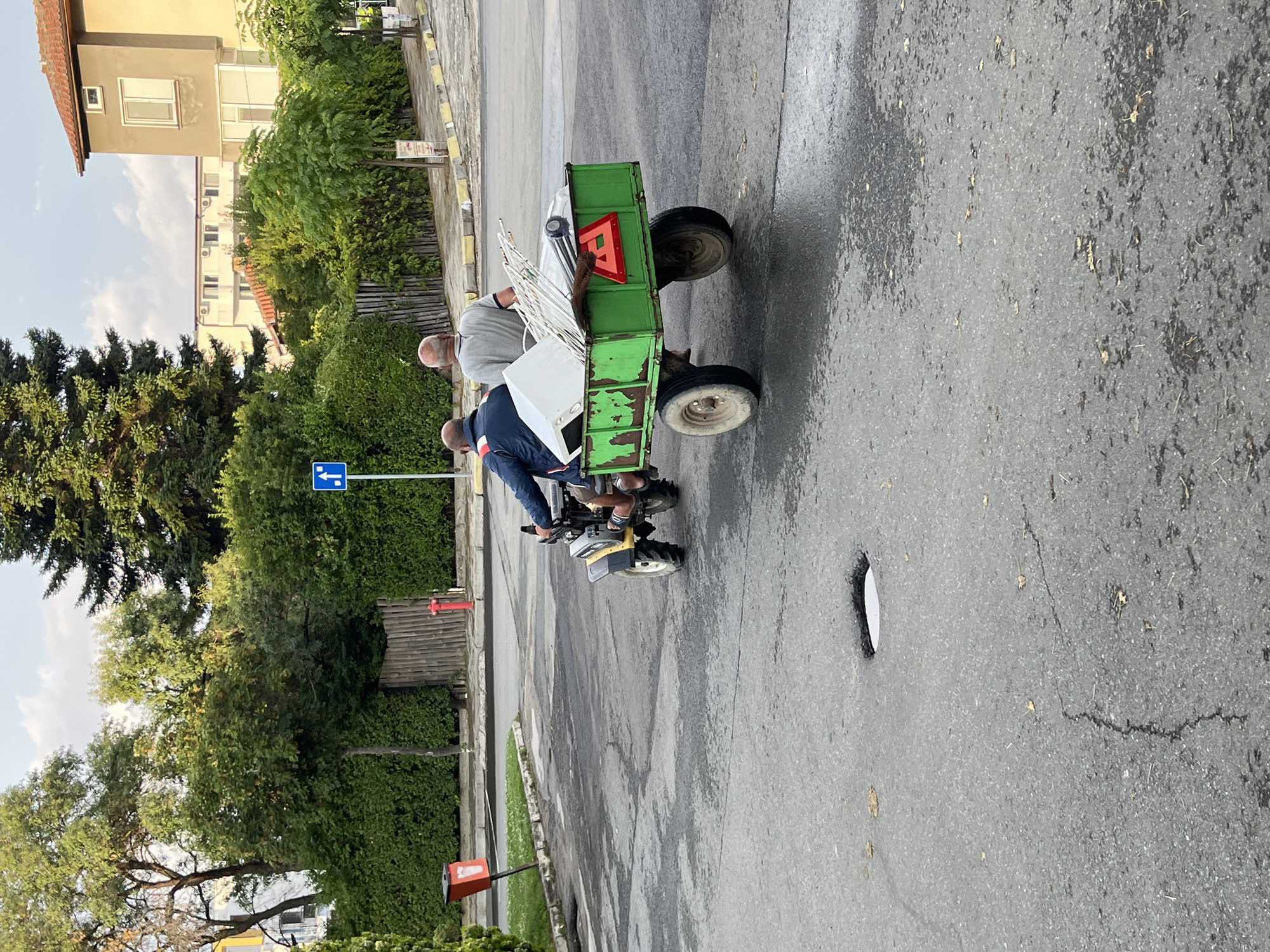 In the city of Obzor, Bulgaria, a small yellow two-wheeled tractor, driven by a man wearing a blue jacket and shorts, pulls a green trailer laden with scrap metal and what appears to be a refrigerator. A second man, wearing a grey shirt, sits in the trailer. They are traveling on a wet asphalt road with potholes and cracks. The surrounding environment includes lush green trees and bushes, a red fire hydrant, a blue street sign indicating a one-way street with a left turn, and residential buildings in the background. The scene is captured from a rear-side perspective, suggesting the subjects are moving away from the camera.